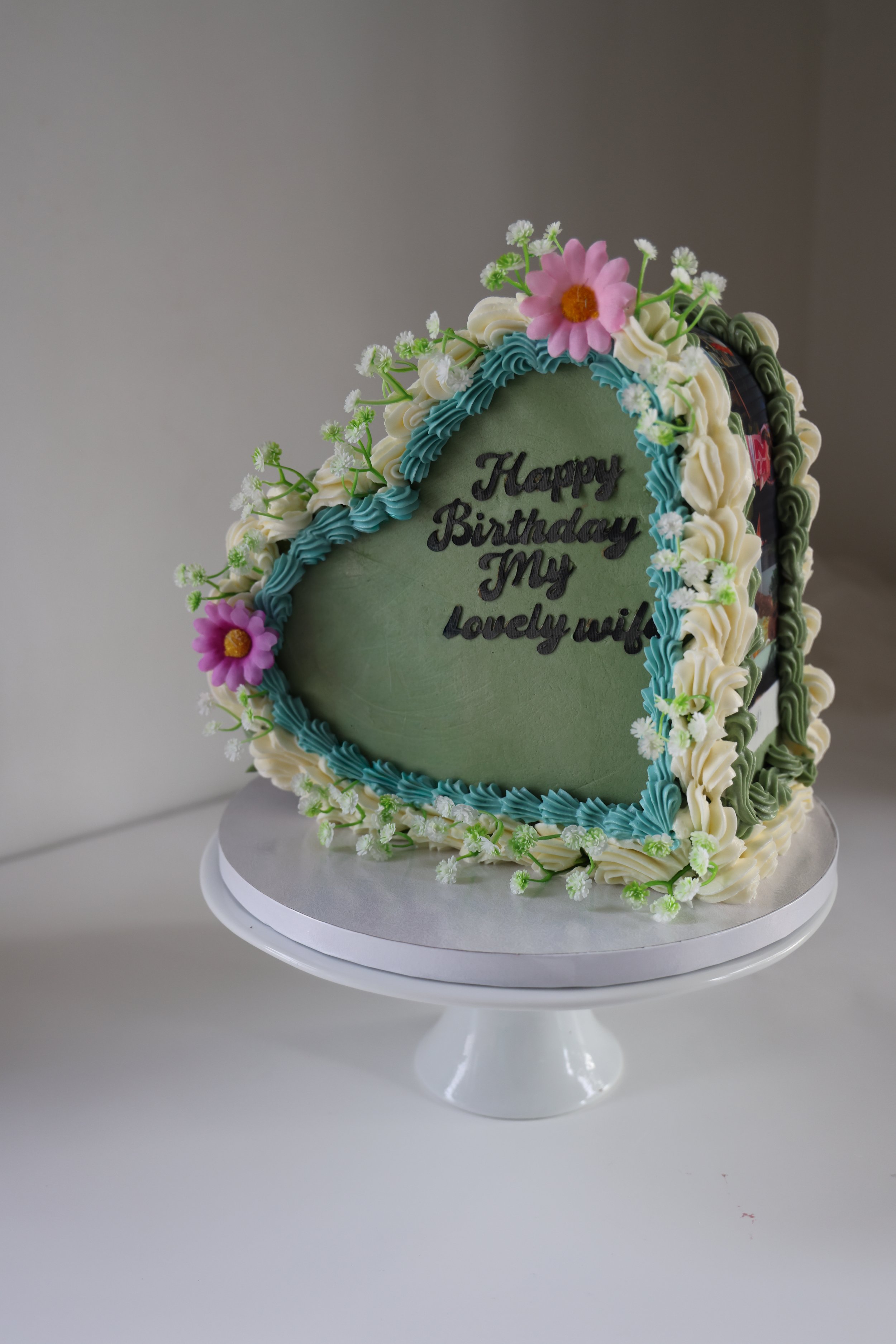 Top sided Heart-shaped birthday cake with flowers and the message "Happy Birthday My lovely wife" in blue icing and edible pictures around the cake.