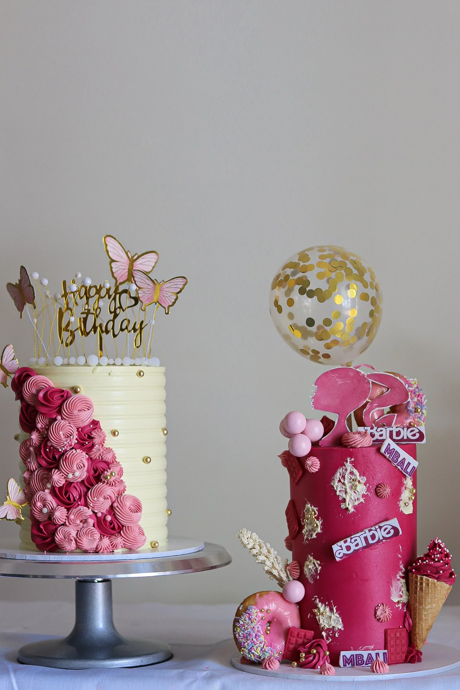 Two decorated Barbie birthday cakes, one tall with pink flowers and butterflies, the other shorter with pink frosting, swan toppers, and ice cream cone decorations, on a table with a balloon in the background. Perfect for girls, pink barbie cake.