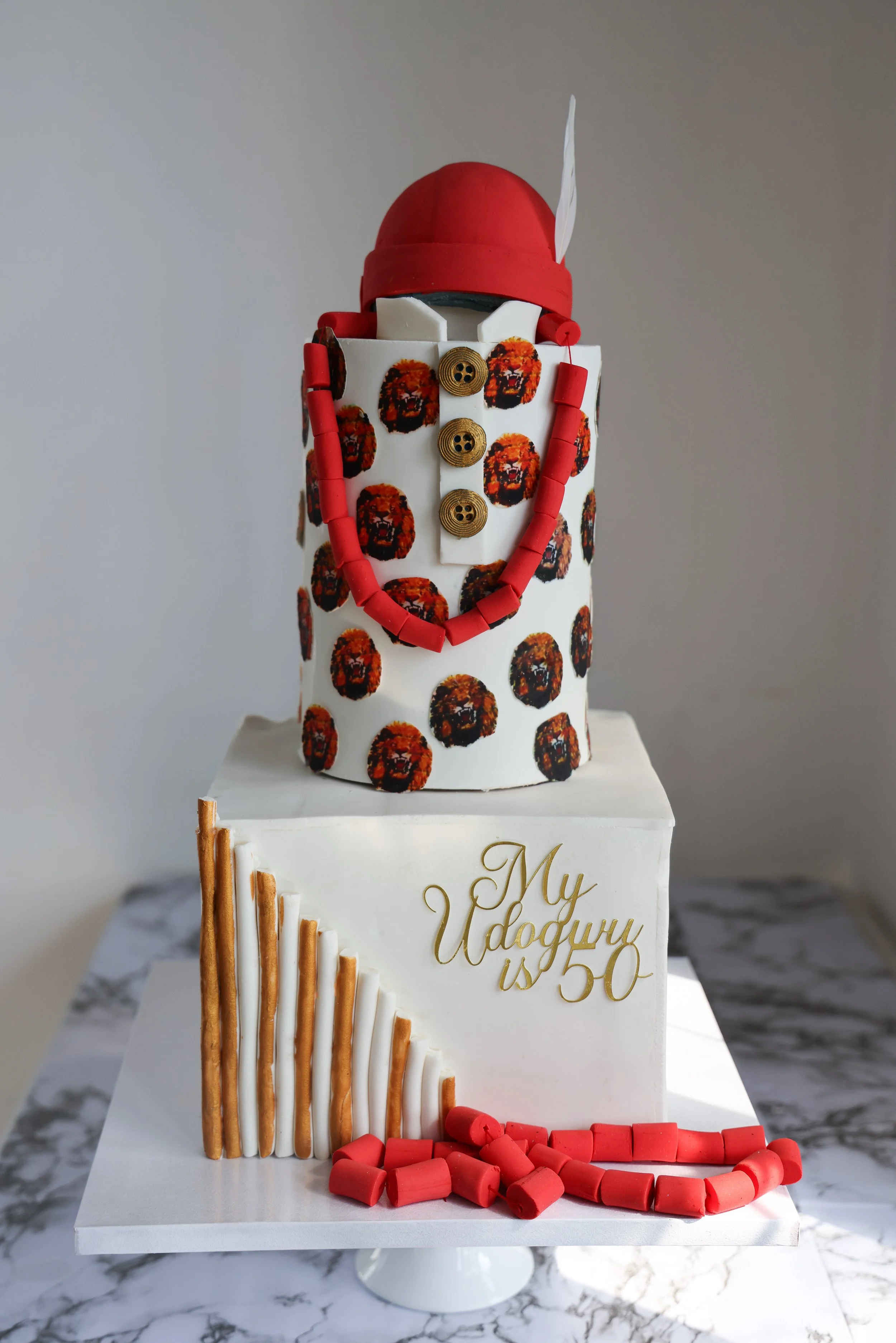 A multi-tiered cake with a Nutcracker soldier theme, featuring a white base with a pattern of tiger faces, red and white beads, gold buttons, and a red hat resembling a Nutcracker's uniform. The cake has a message in gold that reads "My Udobnyi is 50