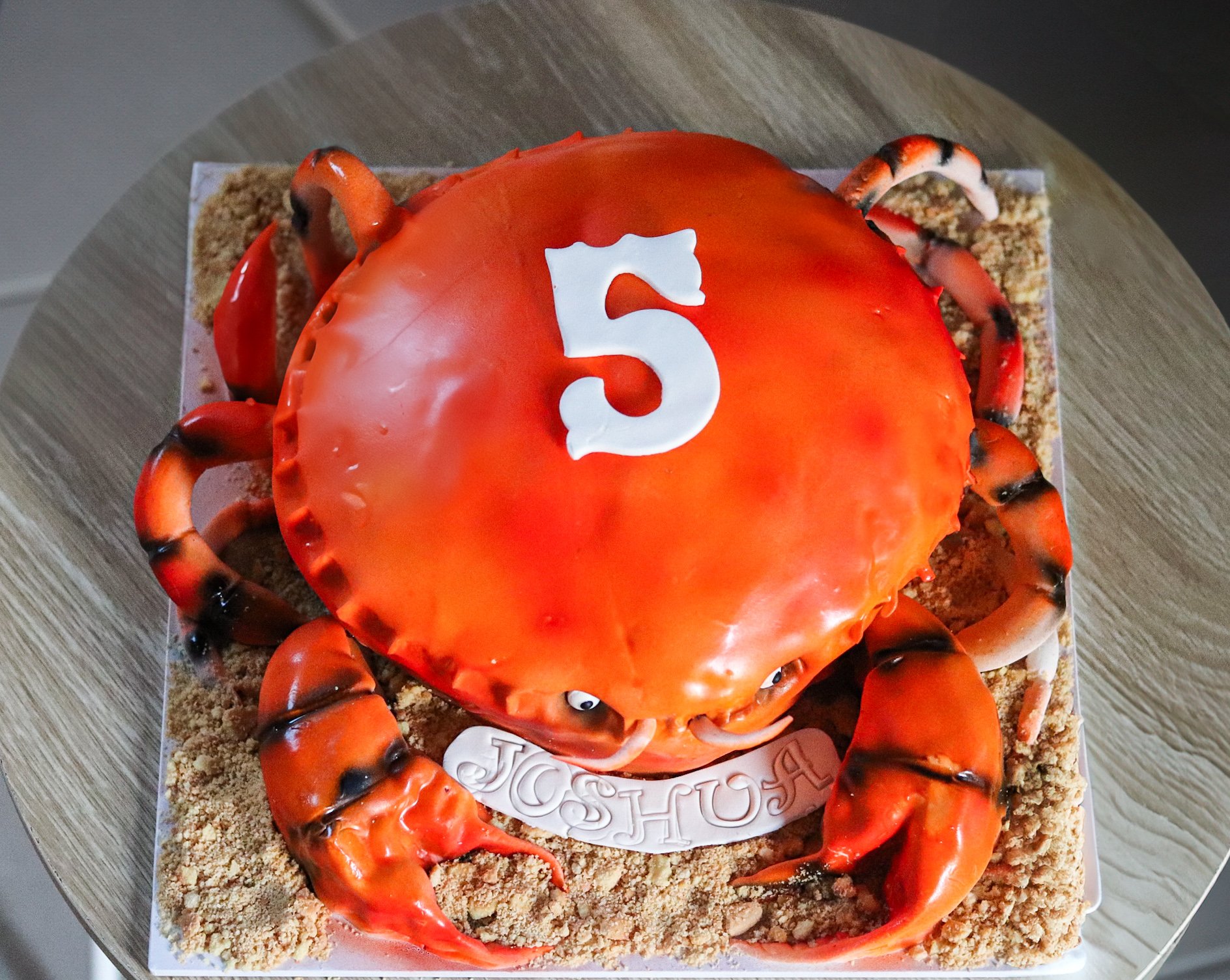 A 3D cake designed to look like a crab, with an orange shell on top featuring a white number 5, surrounded by crab legs and sand-like decorations.