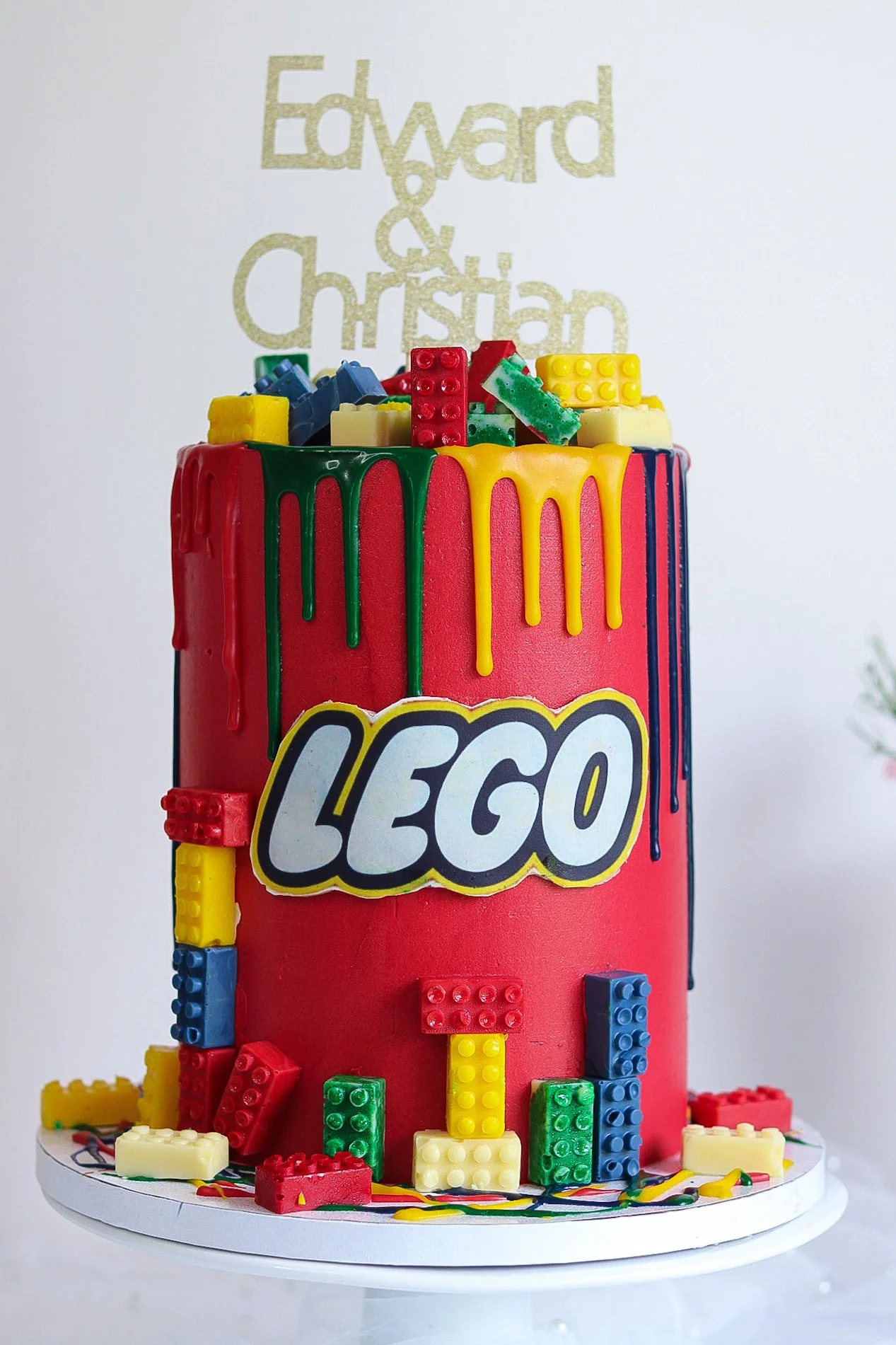 A cake decorated with LEGO bricks and the LEGO logo, with red, yellow, green, and blue icing and toppings, on a white base, with a white background and a faint gold text reading "Edward & Christian."