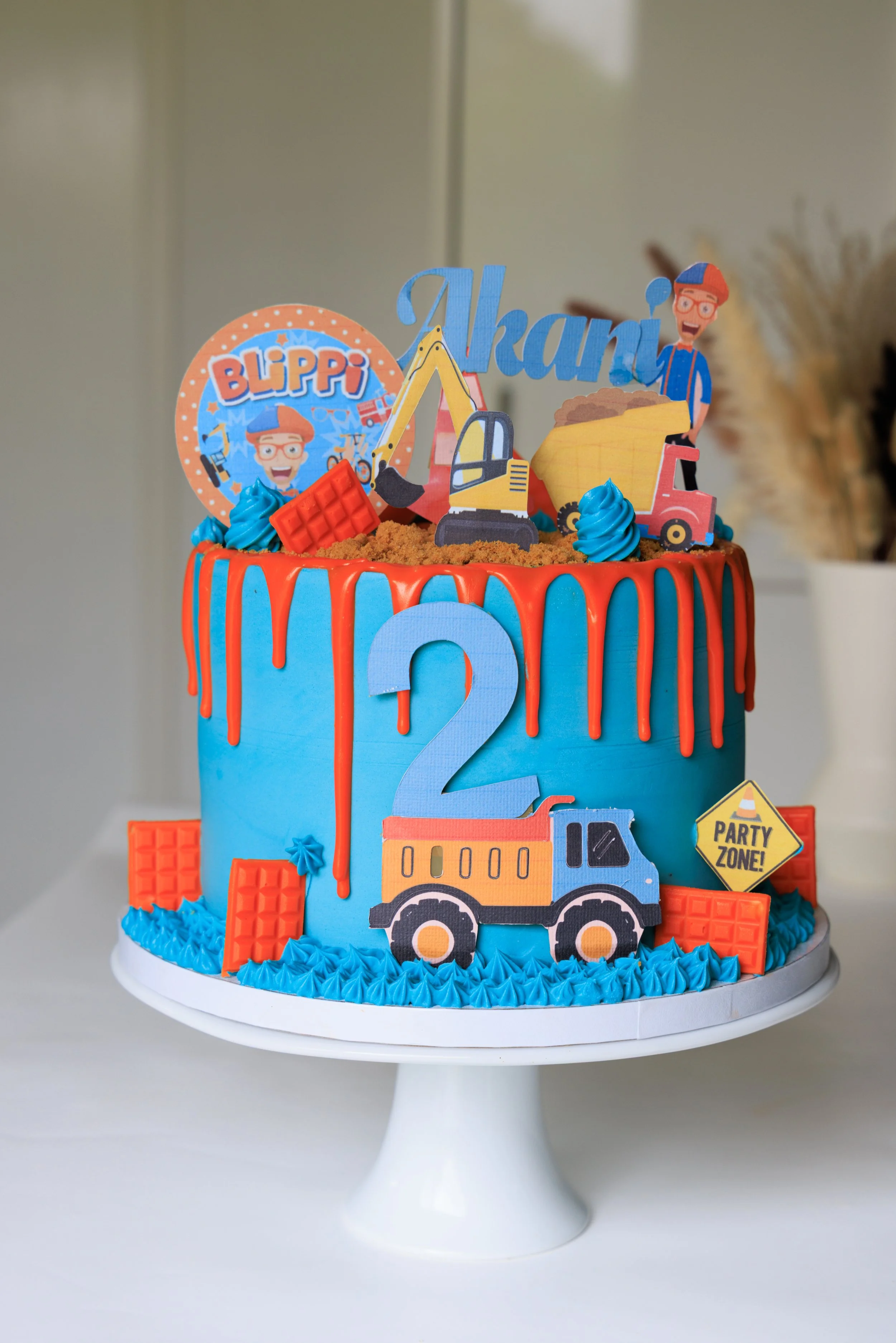 A colorful construction-themed birthday cake with blue frosting, red drip decoration, construction vehicle toppers, a large number 2, and themed decorations including a sign that says "Party Zone".