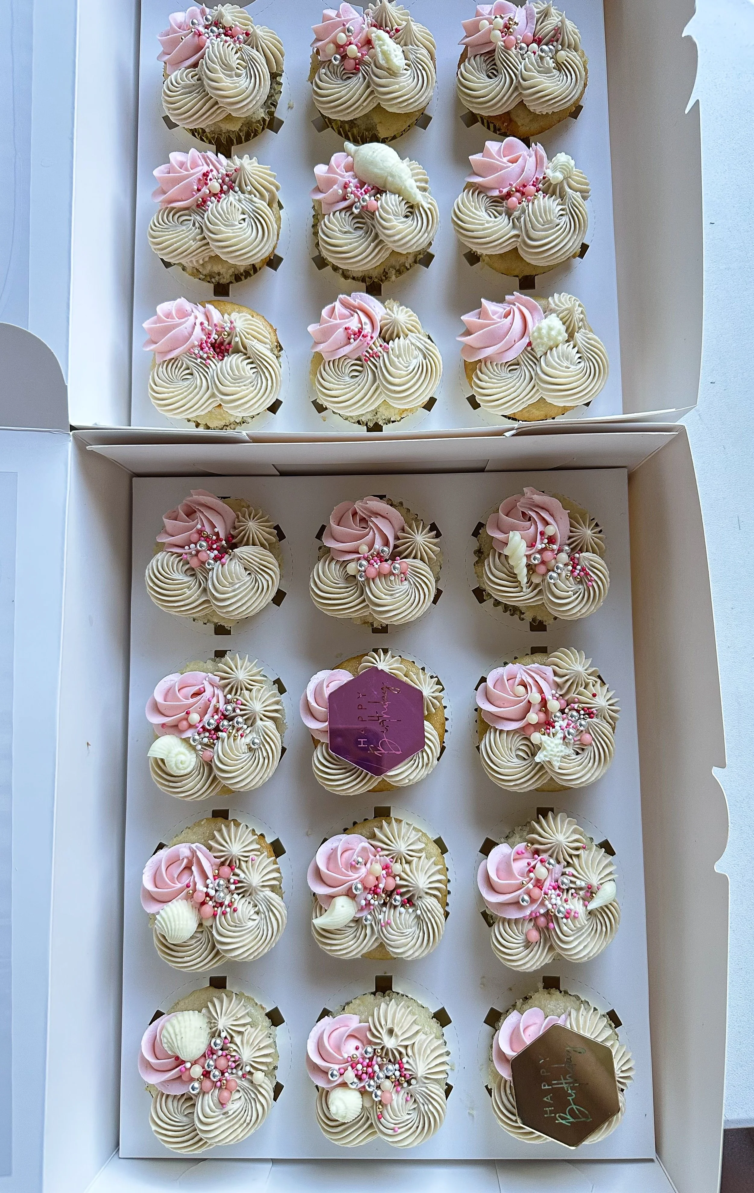 Two white boxes filled with decorated cupcakes. The cupcakes have beige and pink frosting with various pink and white sprinkles and decorations. One cupcake has a small purple 'Happy Birthday' sign on top.