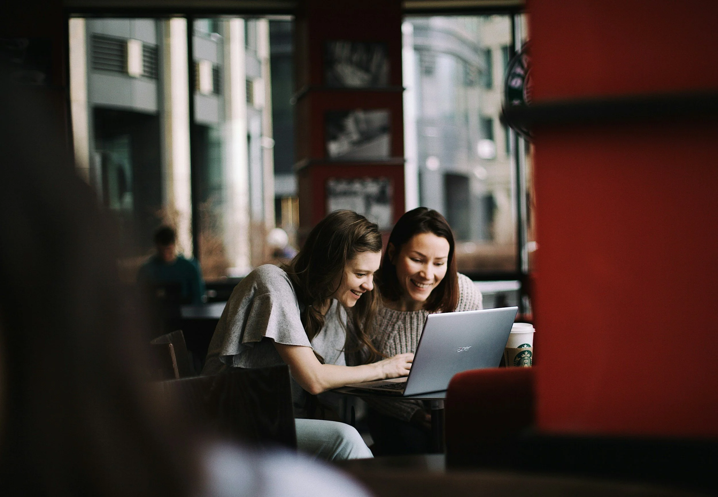 Two women sitting at a table in a coffee shop, looking at a laptop and smiling.