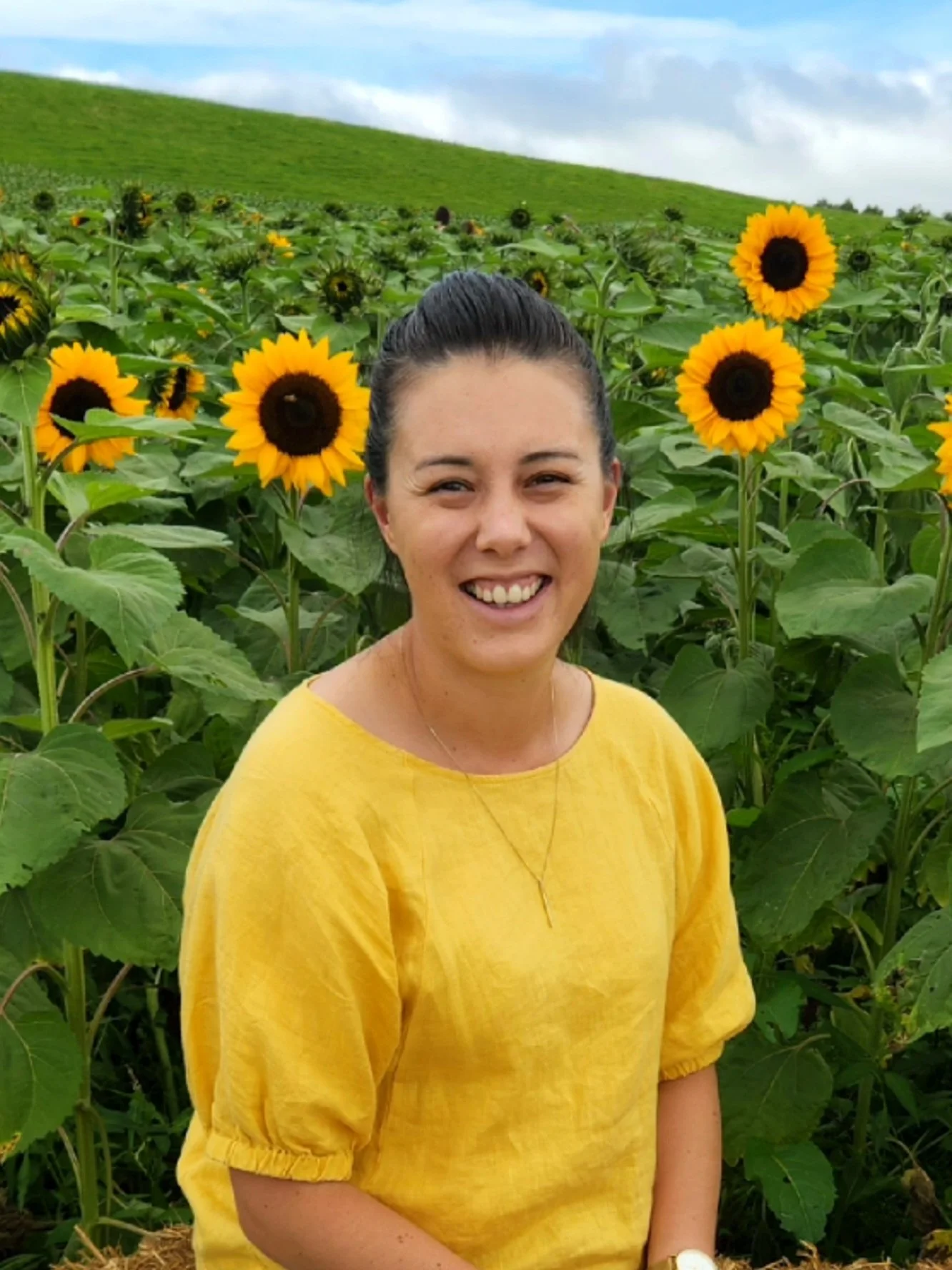 A woman in a yellow top smiling in a sunflower field with green hills and an overcast sky in the background.