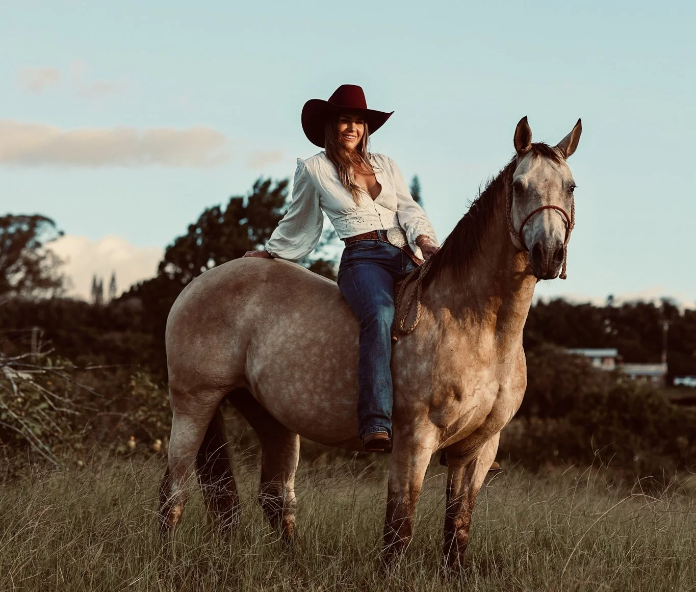 A woman in a white blouse and jeans riding a light-colored horse in a grassy field during sunset.