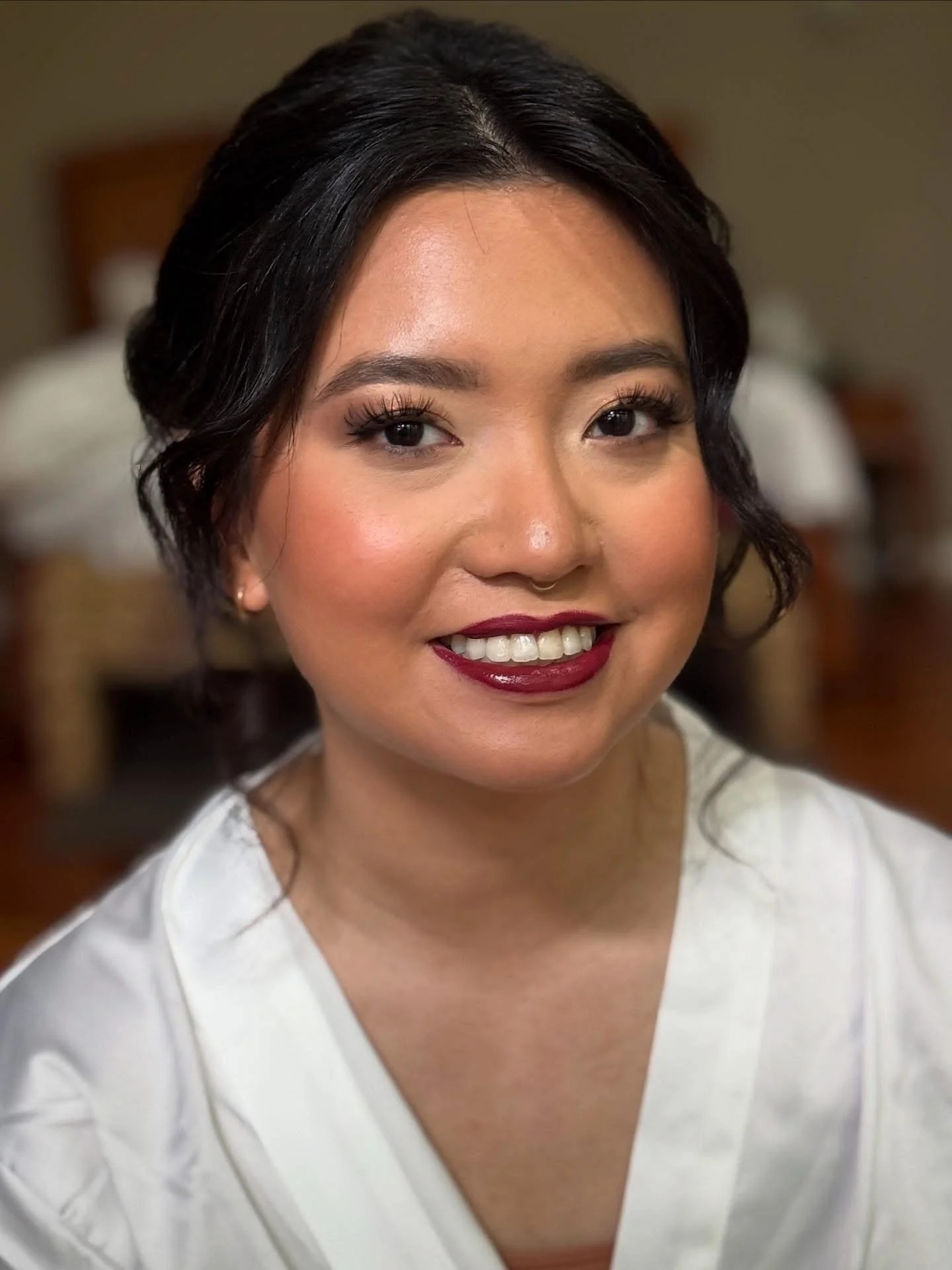 A woman with dark, wavy, shoulder-length hair, wearing makeup with bold lipstick, and a white top, smiling at the camera in a softly lit room.
