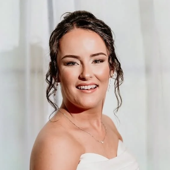 Young woman with dark, curly hair styled in loose waves, smiling and wearing a white strapless dress with a delicate necklace, in a bright room with white curtains.