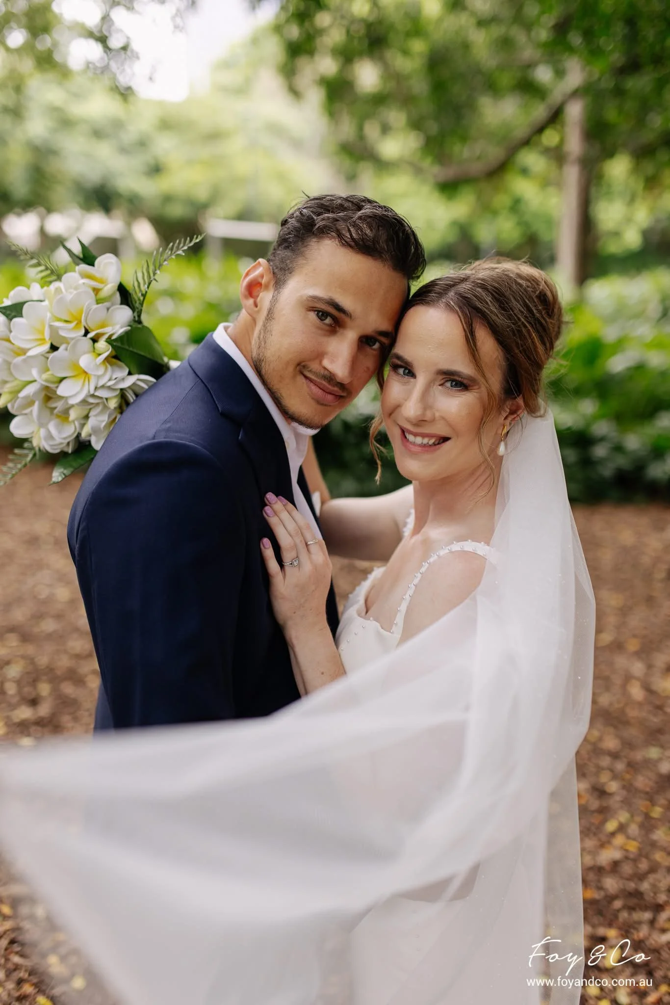 A newlywed couple in wedding attire smiling and embracing in a lush outdoor setting, with the bride holding a bouquet of white flowers.