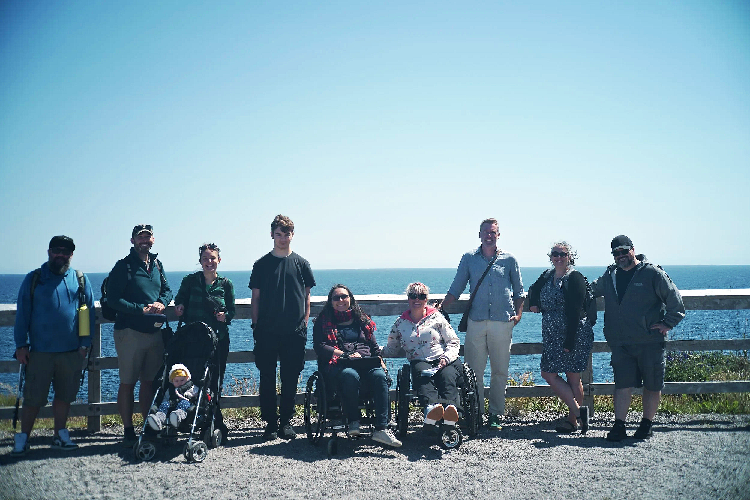 A group of ten people, including children in strollers, standing on a gravel path by a wooden fence with ocean in the background on a sunny day.