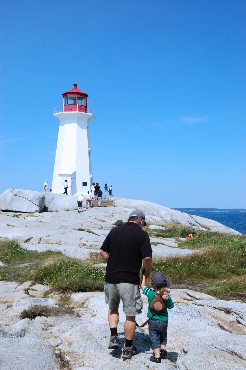 A lighthouse on rocky terrain with multiple visitors around it, and an adult and child walking hand in hand towards the lighthouse.