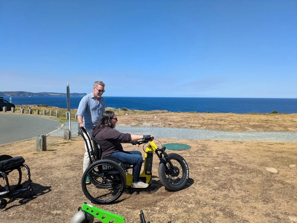 A woman in a wheelchair riding an electric bicycle along a coastal area with two men nearby, one standing and one sitting.