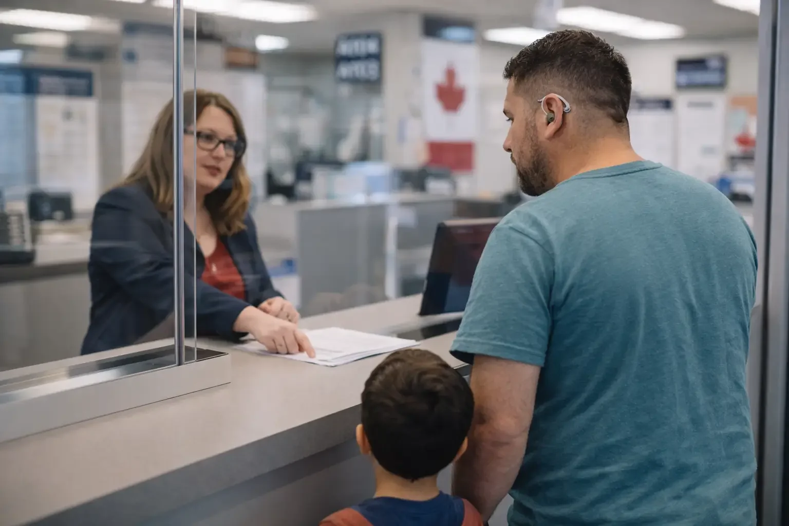 A man and a child standing at a bank teller window, talking to a woman behind the glass.