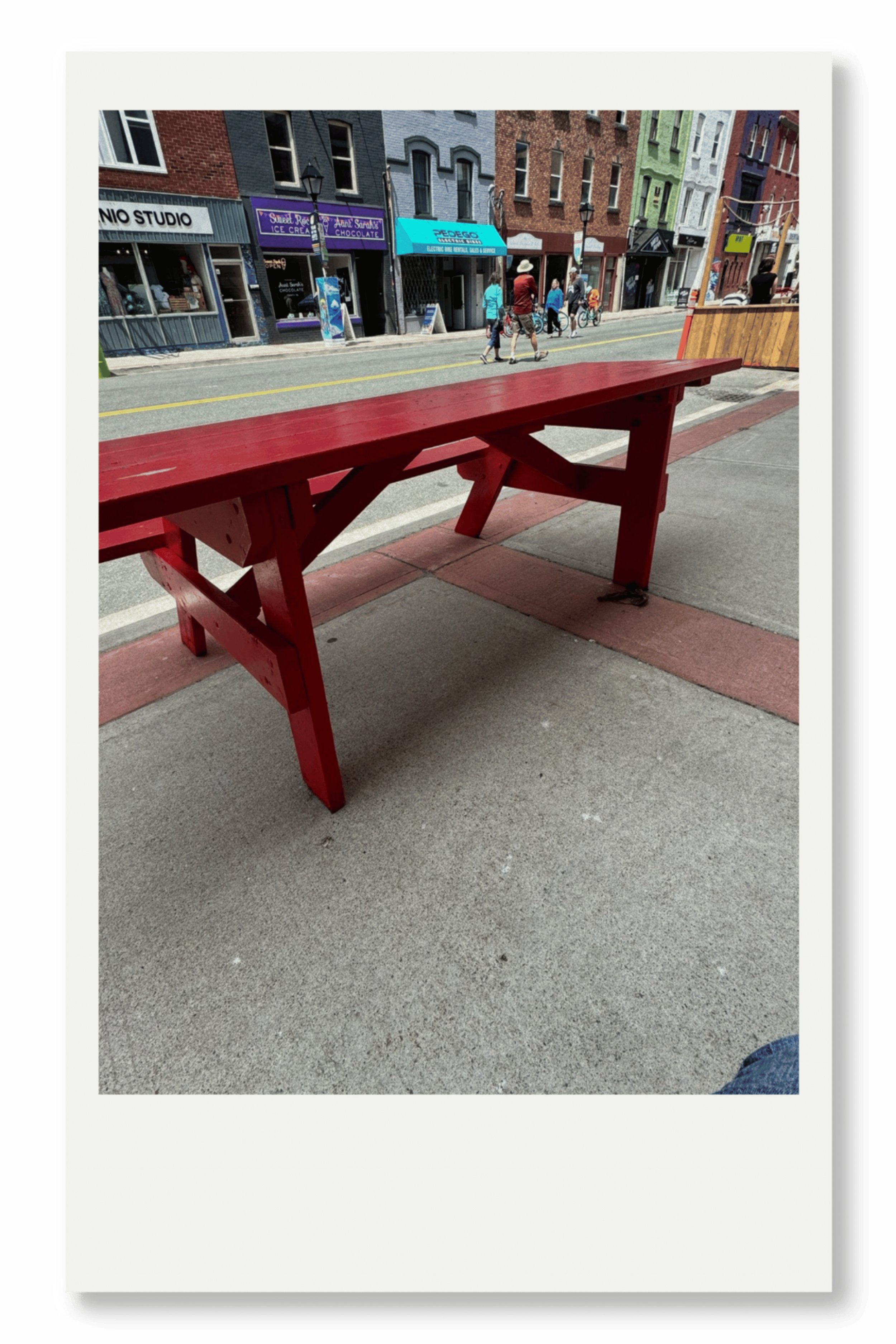 A red wooden bench on a sidewalk in front of a colorful city street with shops and pedestrians.