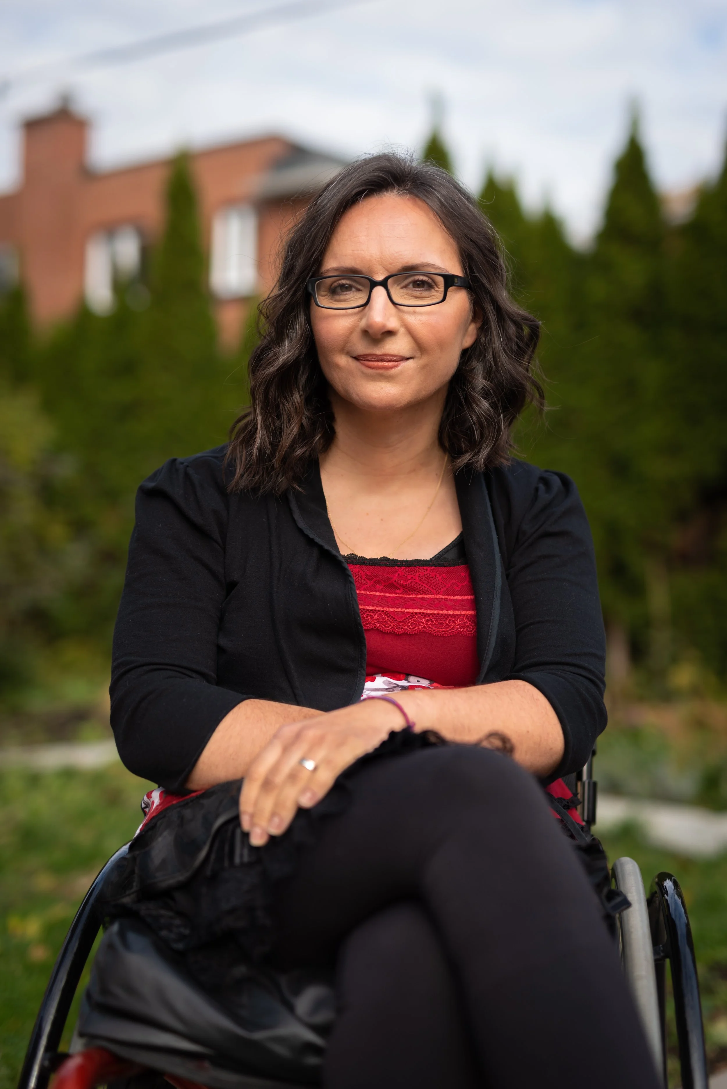 Dr. Marjorie Aunos with shoulder-length wavy dark hair, wearing black glasses, a black cardigan, a red top with lace, and black pants, sitting outdoors in a garden or park with trees and a house in the background.