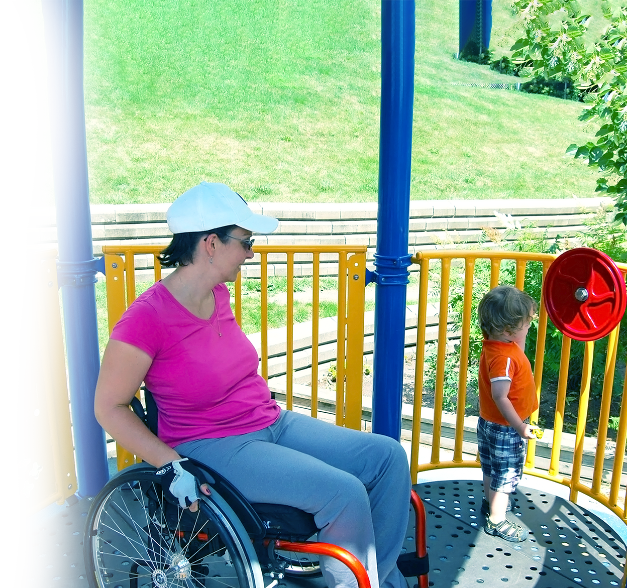 A woman in a wheelchair and a young boy at a playground. The woman is wearing a pink shirt, gray pants, sunglasses, and a white cap. The boy is wearing an orange shirt and plaid shorts. They are near a yellow and blue play structure with a red steering wheel, set outdoors on a sunny day.