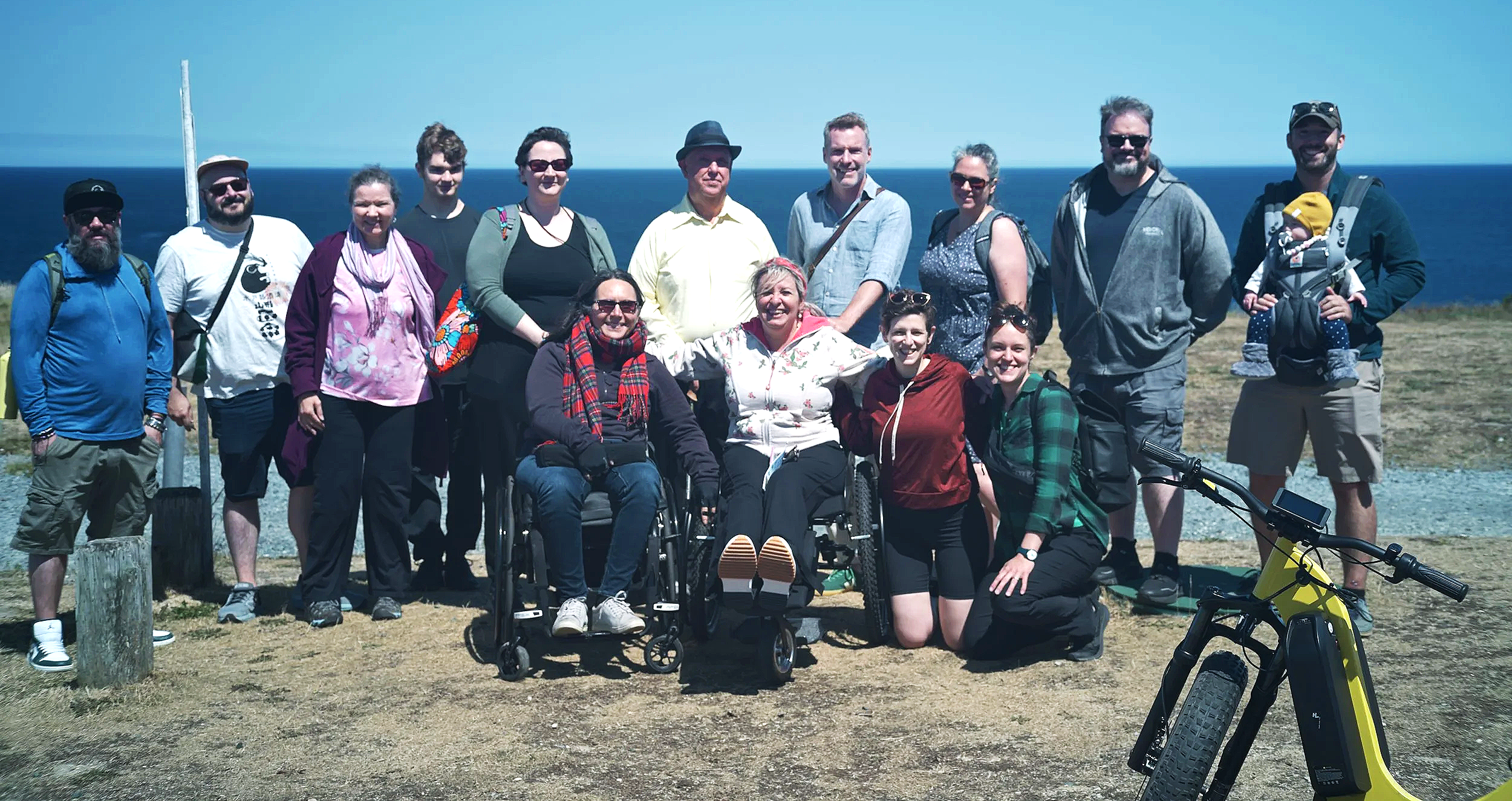 A group of people outdoors on a sunny day, with the ocean in the background. Some are standing, others are seated in wheelchairs, and one person holds a baby. A bicycle is visible in the foreground.