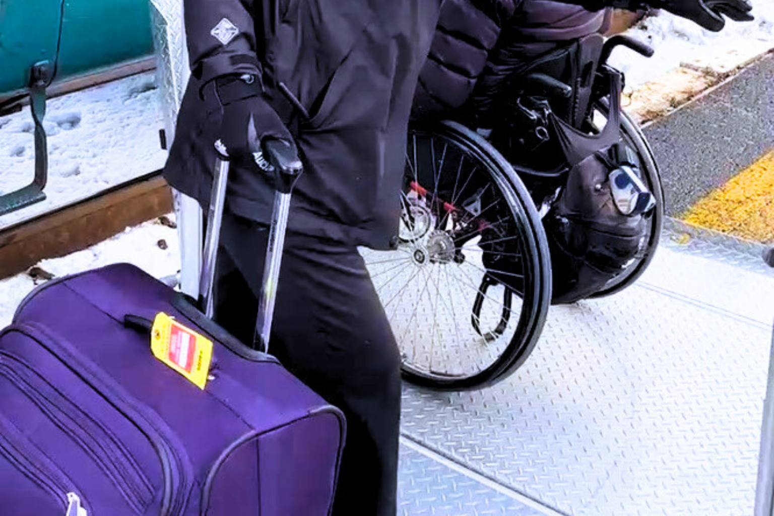Person with a purple rolling suitcase and a wheelchair with a bag on it, standing on a metal boarding platform with snow on the ground.