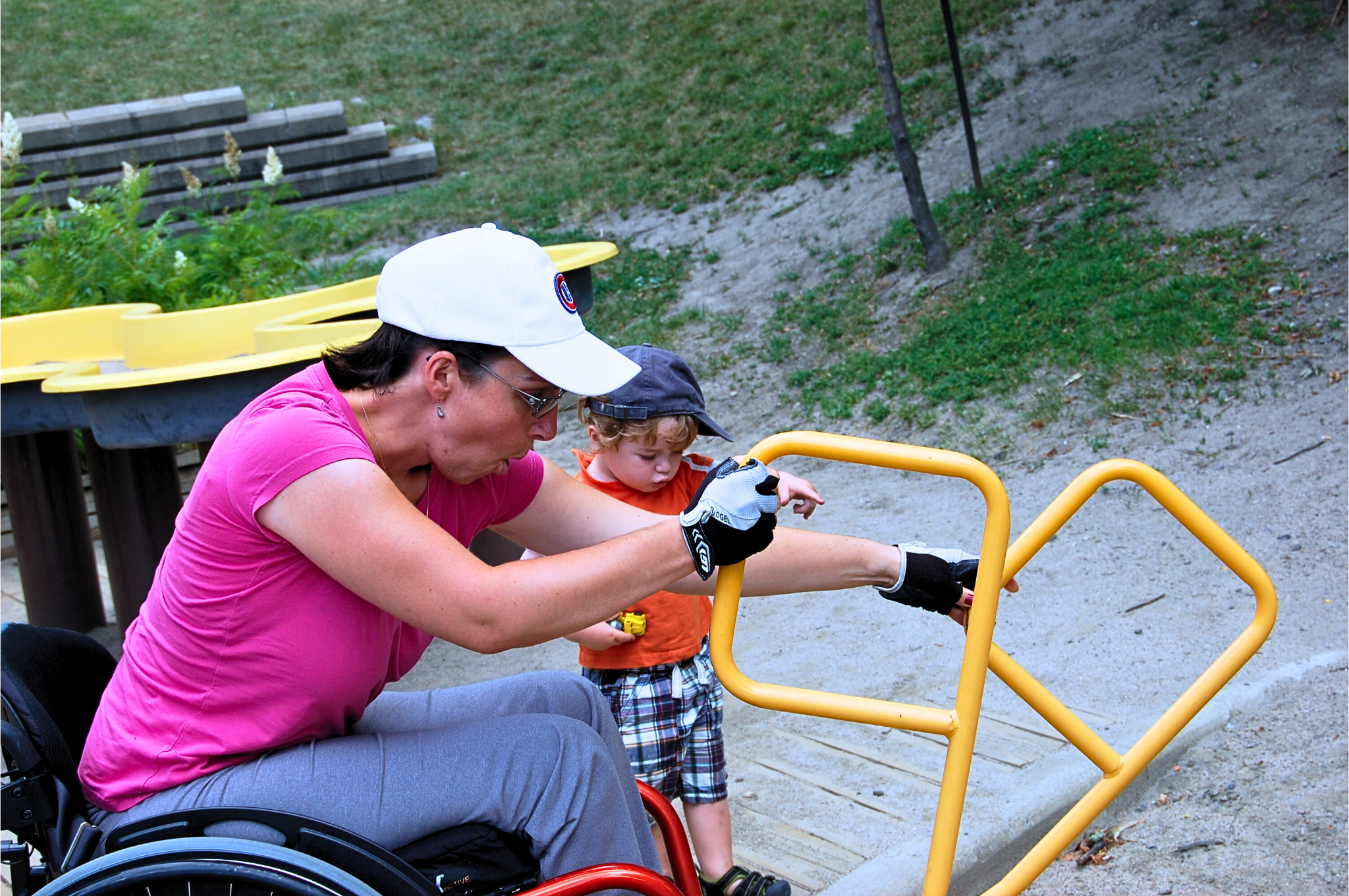 A woman in a pink shirt and white cap helping a young boy with a disability with a wheelchair by holding a yellow handrail on a playground. The boy is reaching out to hold the yellow handrail.