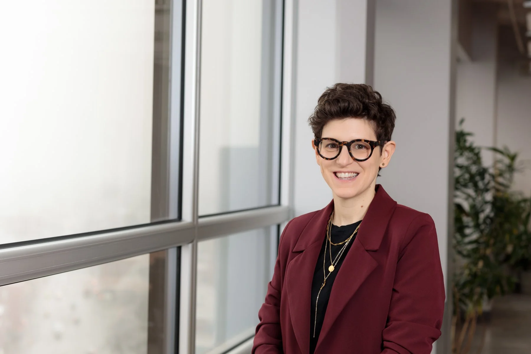 Dr. Coralie Mercerat, short dark hair, glasses, and a maroon blazer standing next to large windows in a modern office, smiling.
