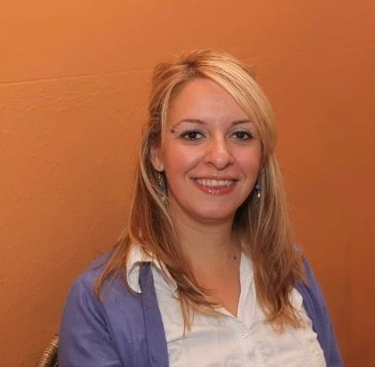 Dr. Laura Pacheco smiling with shoulder-length blonde hair, wearing a white blouse and a purple cardigan, sitting against an orange wall.