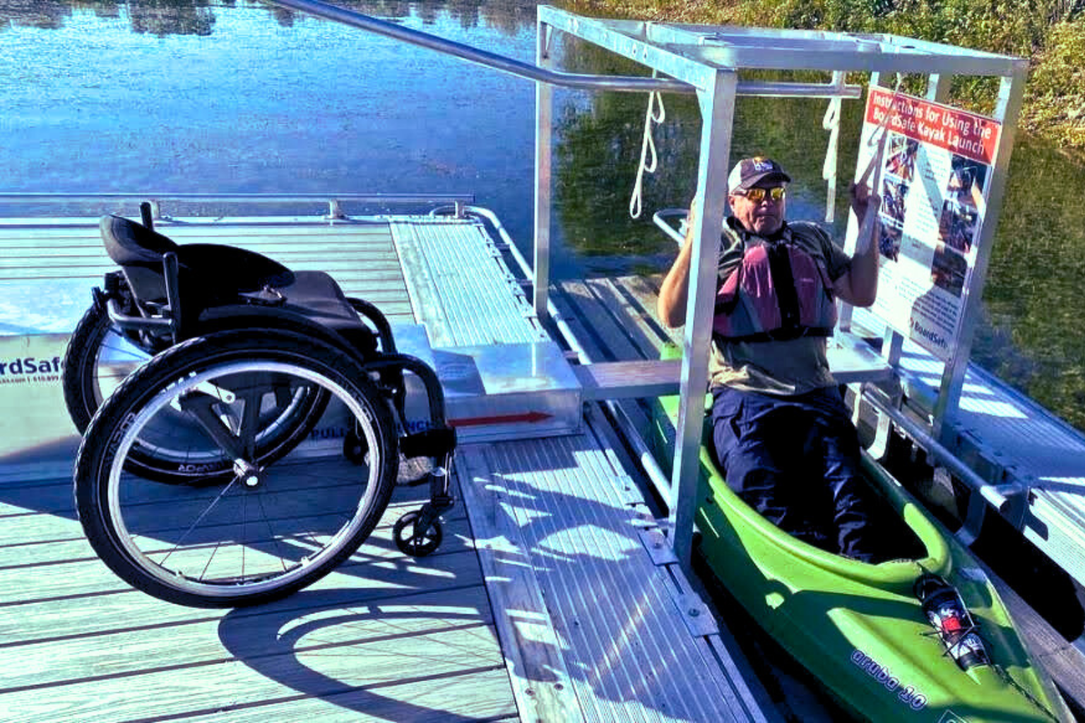 A man sitting in a green kayak on a dock, holding a newspaper, next to a wheelchair, with water and trees in the background.