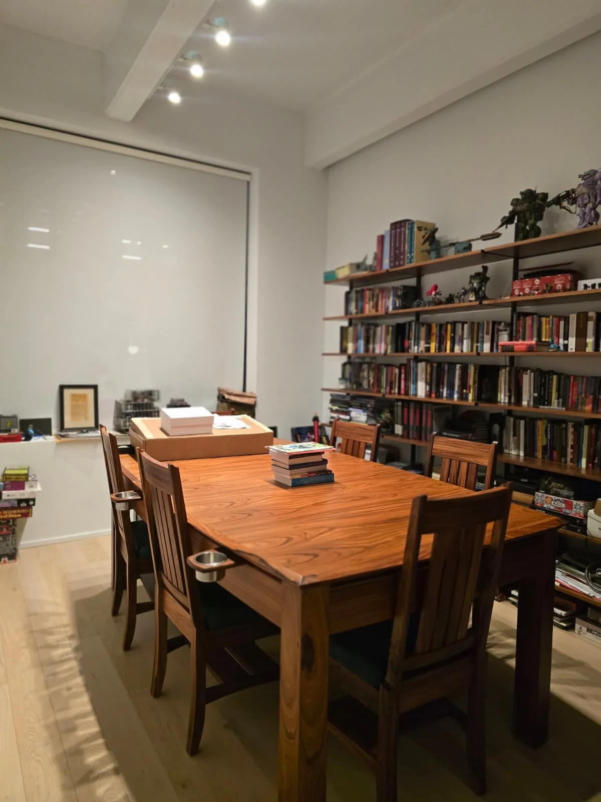 A dining room with a rectangular wooden table, six chairs, a tall bookshelf filled with books and figurines against the wall, and a large window with a white roller shade.