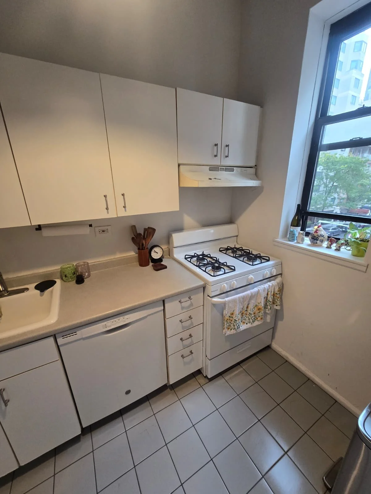 A small kitchen with white cabinets, a white gas stove with a floral towel hanging from its handle, a white dishwasher, and a window with decorative items on the windowsill, including a plant and small figurines.