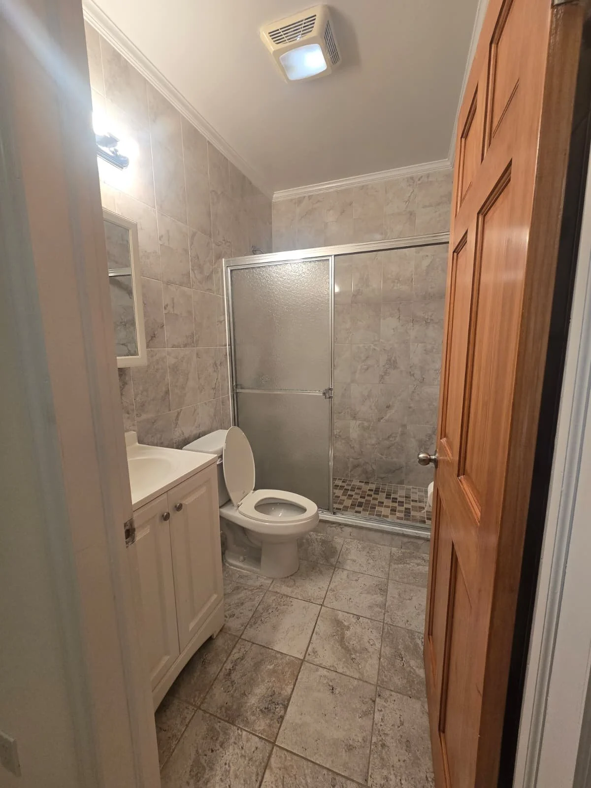 Small bathroom with beige tiled walls and floor, wooden door, white vanity with sink, toilet, and glass shower stall.