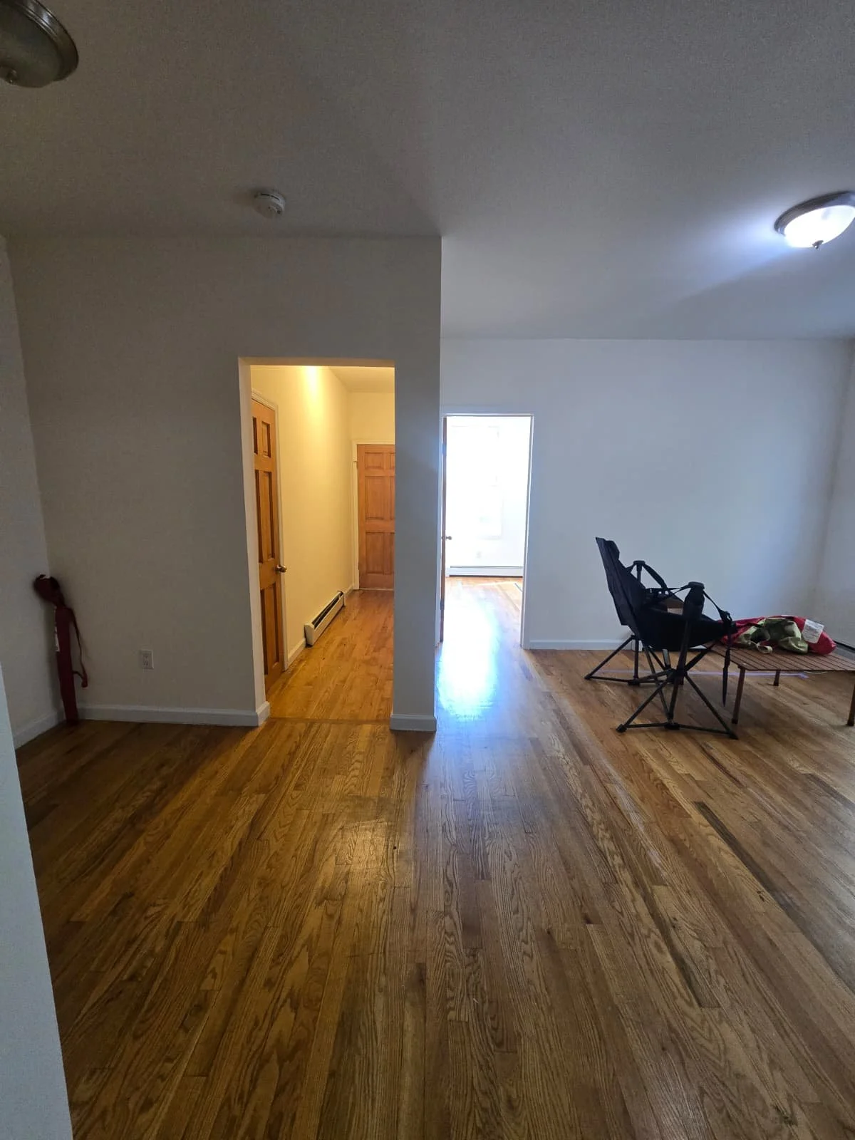 Empty living room with hardwood floors, a black folding chair and a small table with some items on it, and a doorway leading to other rooms.