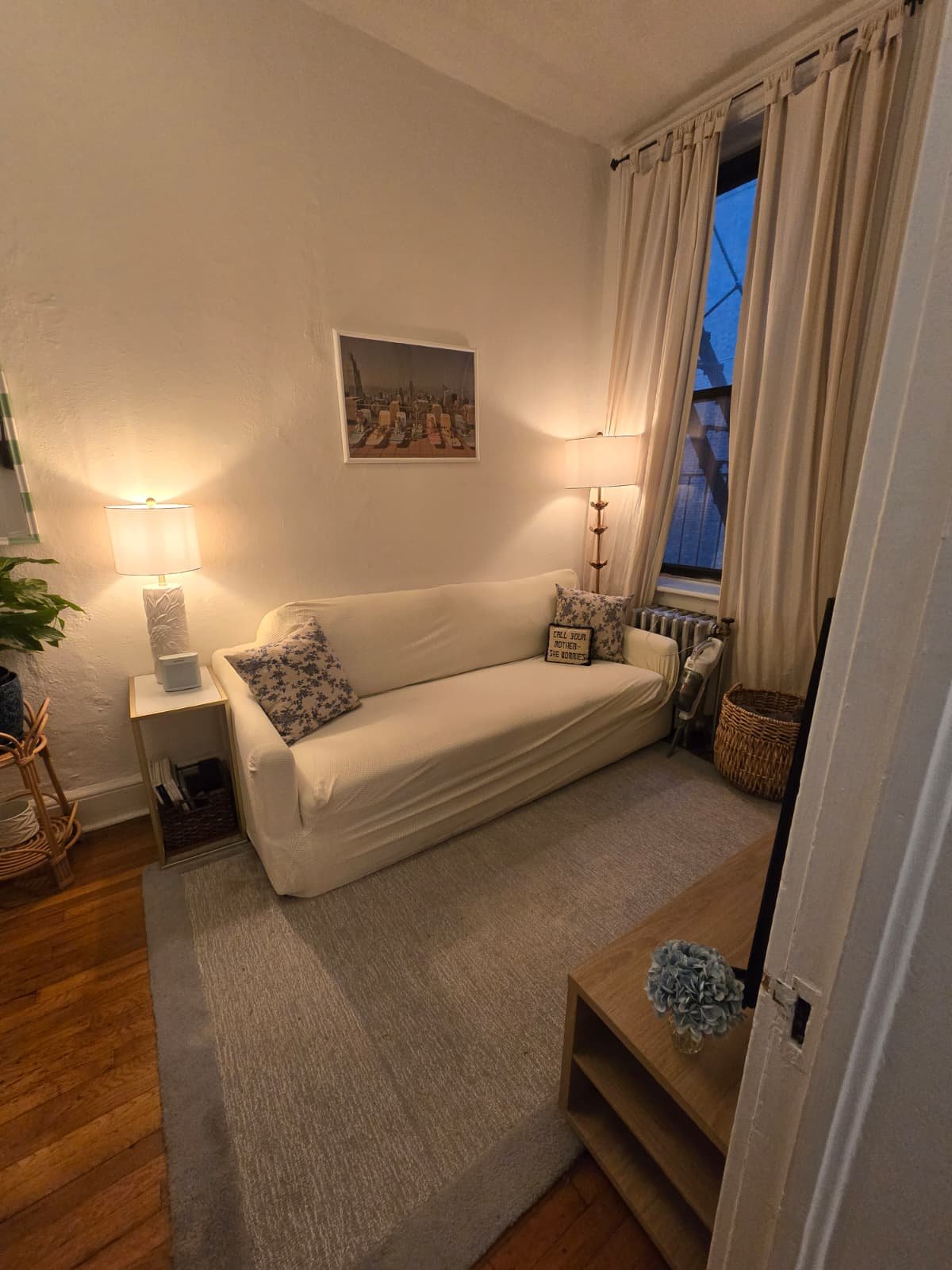 Living room with a white sofa, two lamps, a framed picture on the wall, a window with cream curtains, a wicker basket, and a beige rug on hardwood floor.
