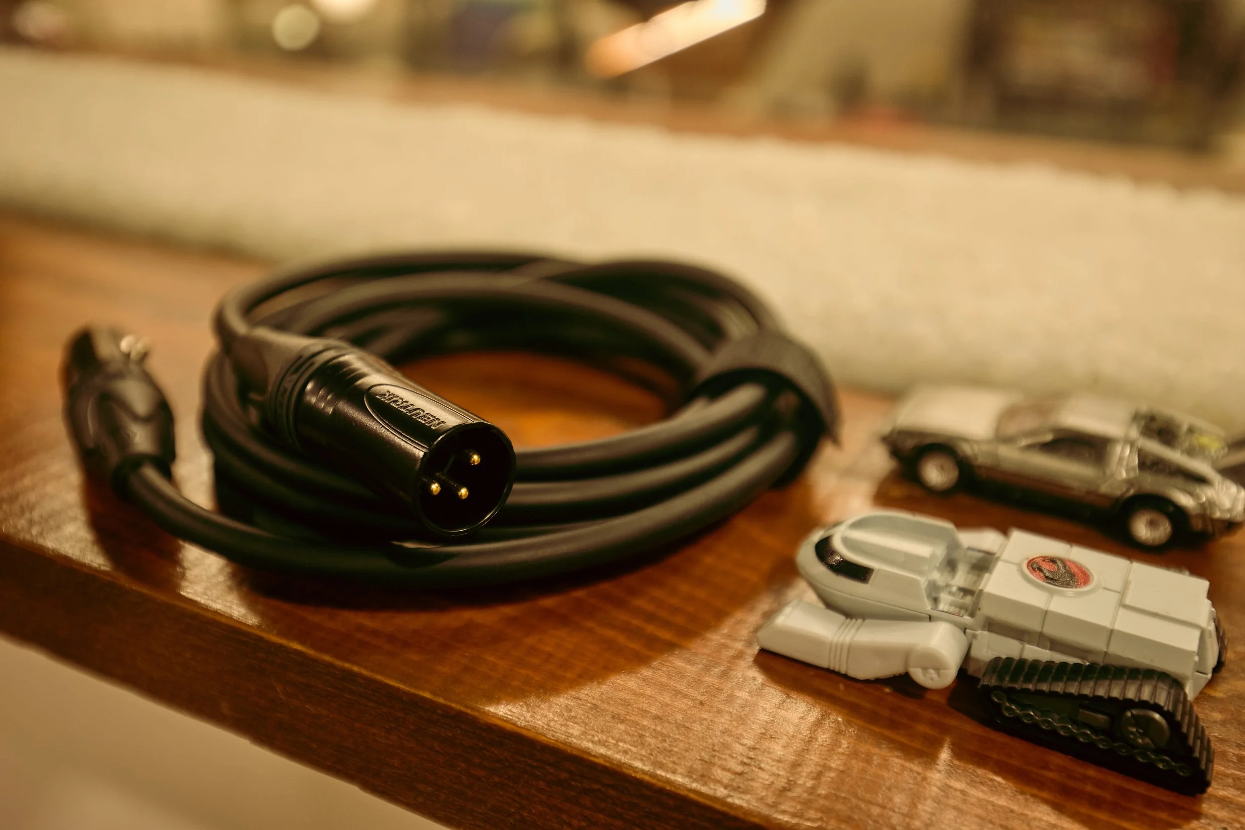 A coiled black power cable with a three-prong plug, two toy cars resembling a silver sports car and a white futuristic vehicle, and a small white toy tank on a wooden surface.