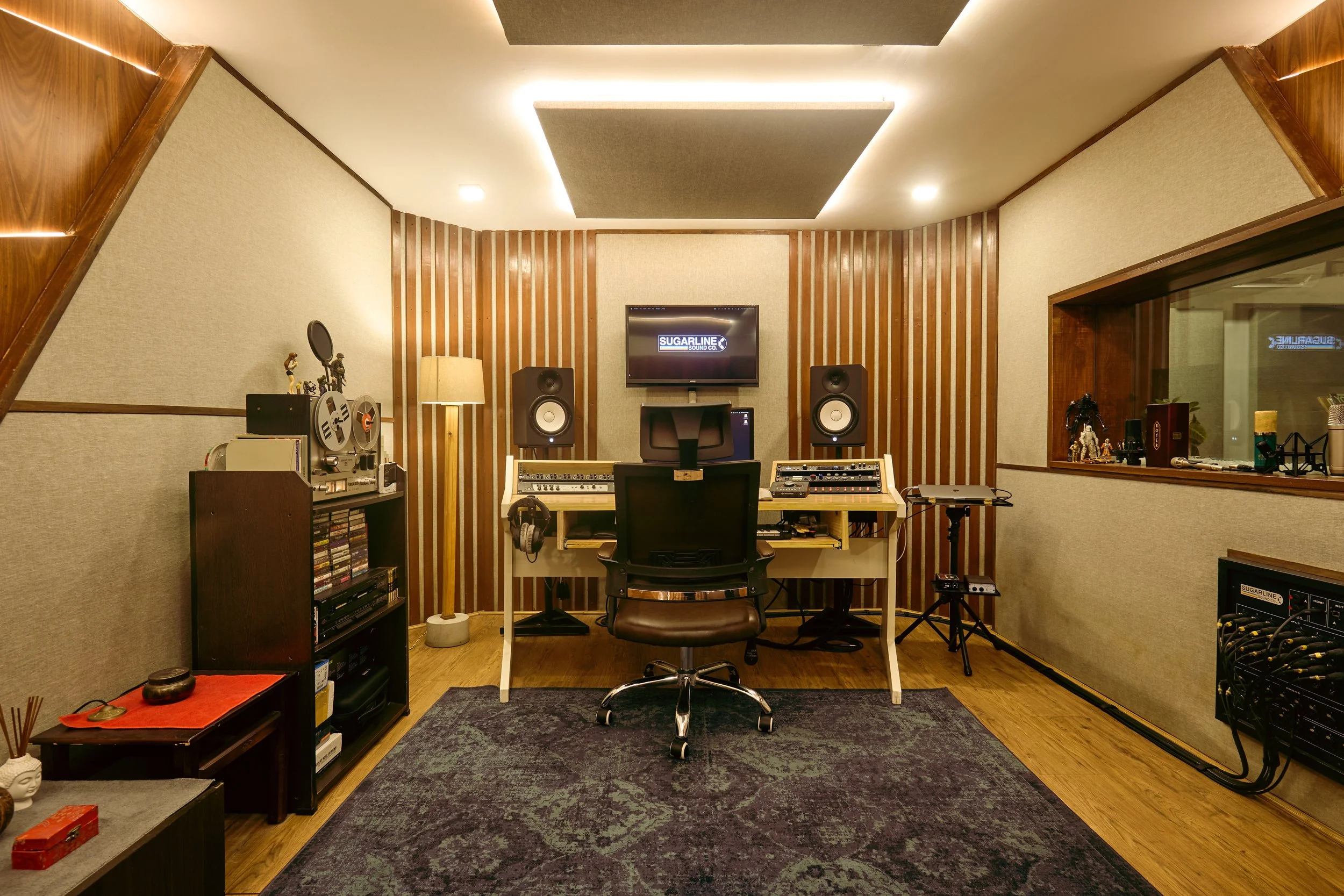 Recording studio with a desk, computer monitor, headphones, and audio equipment arranged in a soundproof room with wood paneling and a carpeted floor.