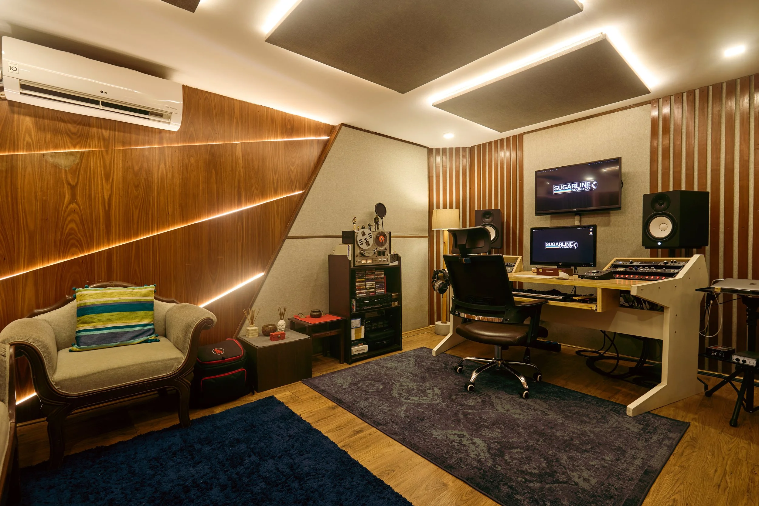 Sound studio with beige and wood paneled walls, a beige vintage sofa with a striped pillow, a blue rug, a black office chair, a wooden desk with musical equipment, and wall-mounted monitors displaying 'SUGARLINE SOUND CO.'.