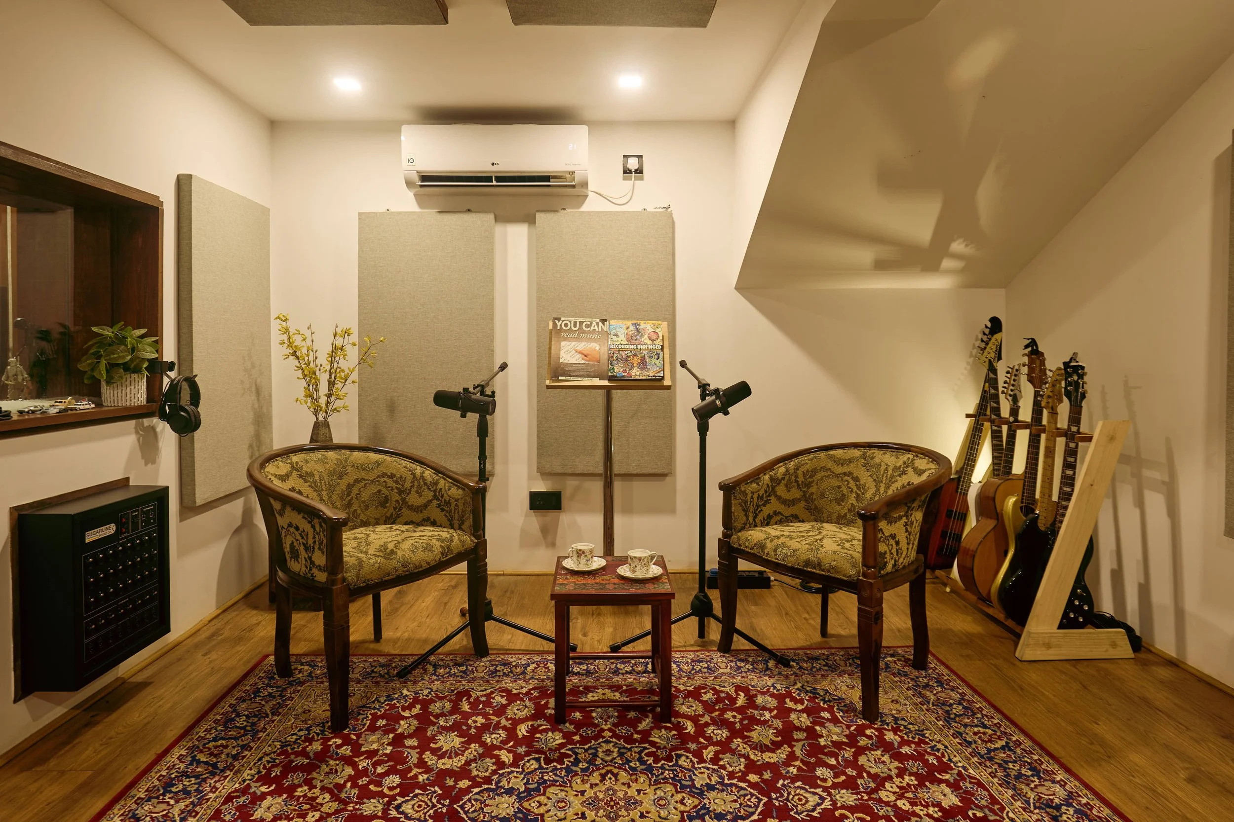 Recording studio with two vintage armchairs, microphones, a small table with two cups, guitar stand with various guitars, soundproofing panels, and musical accessories.