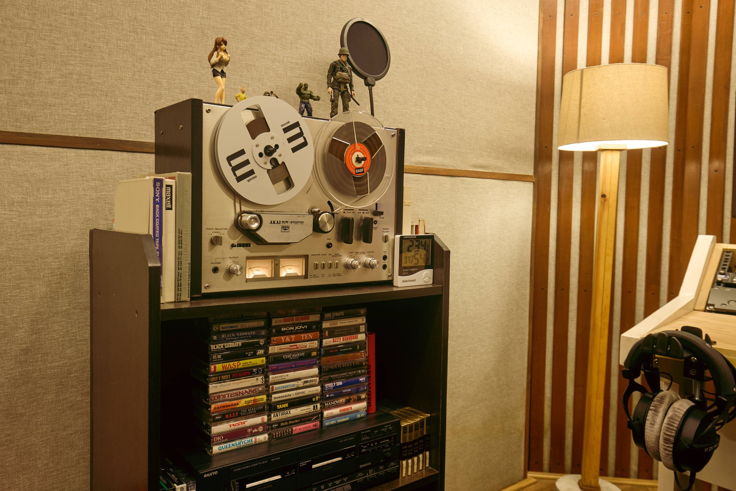 A vintage reel-to-reel tape recorder with figurines on top, a collection of cassette tapes below, and a pair of headphones hanging nearby in a cozy room with wooden paneling and a floor lamp.