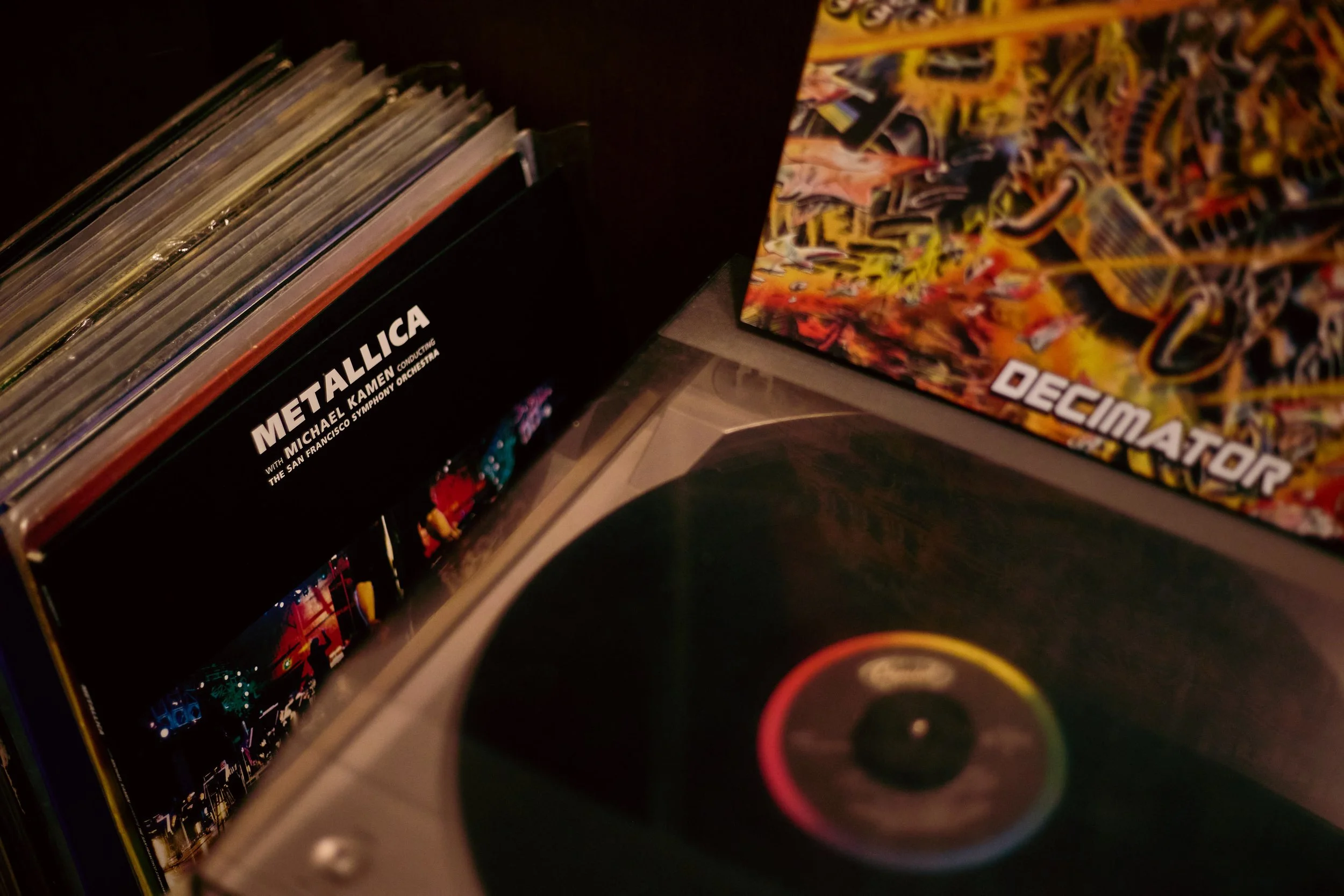 Close-up of a record collection with a Metallica album and a Decimator album, and a turntable with a black vinyl record.