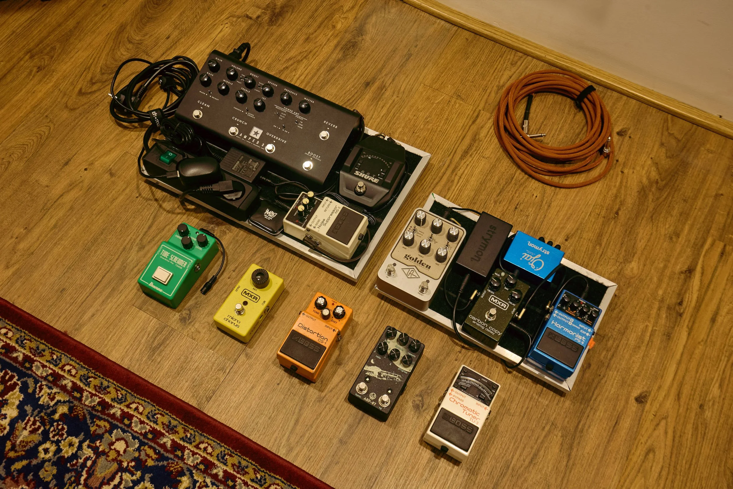 Arrangement of guitar effect pedals and equipment on a wooden floor, including multiple pedals of various colors, a pedalboard, and a coiled orange instrument cable.