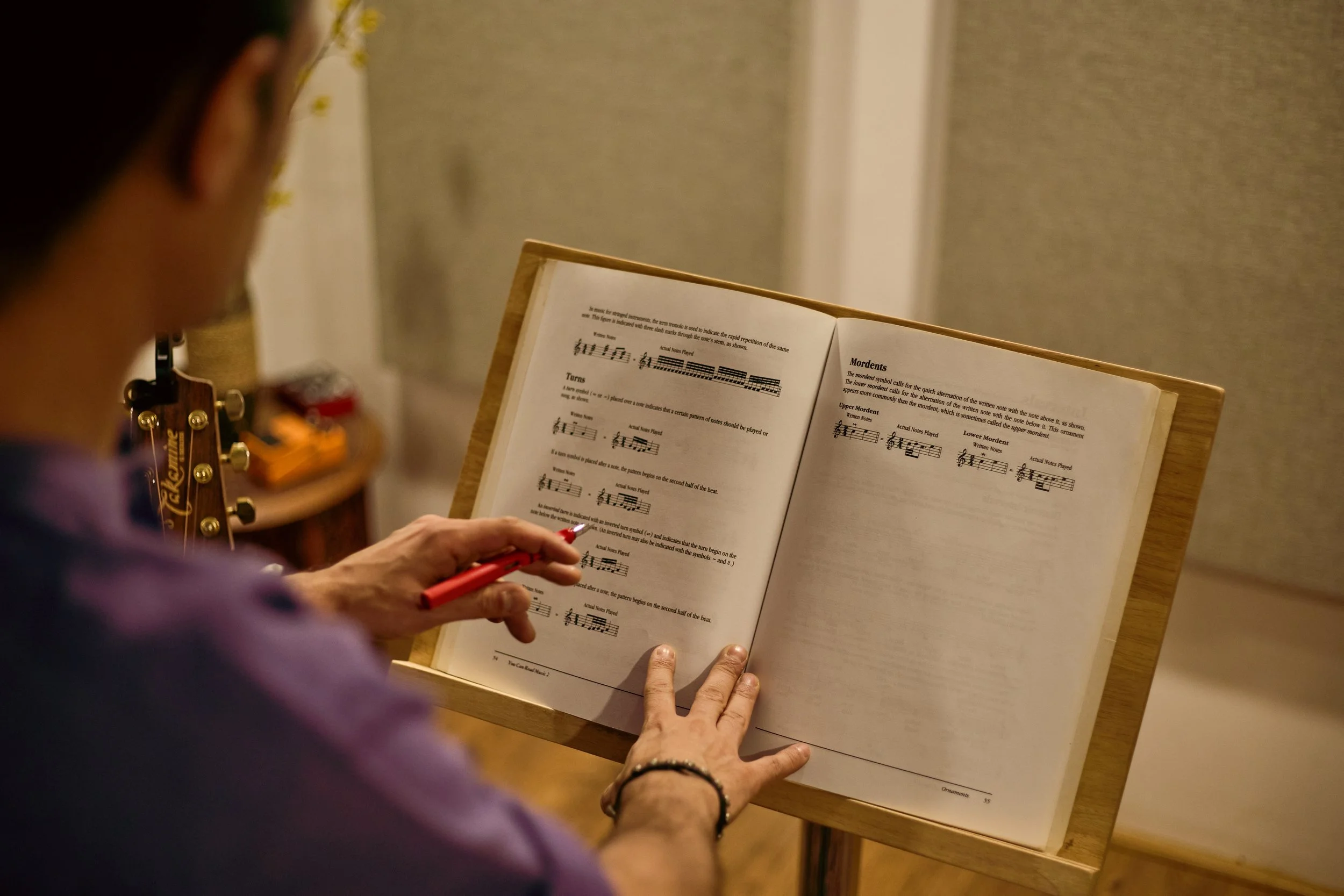 Person holding a red pen and reading sheet music notes on a music stand, with a guitar in the background.