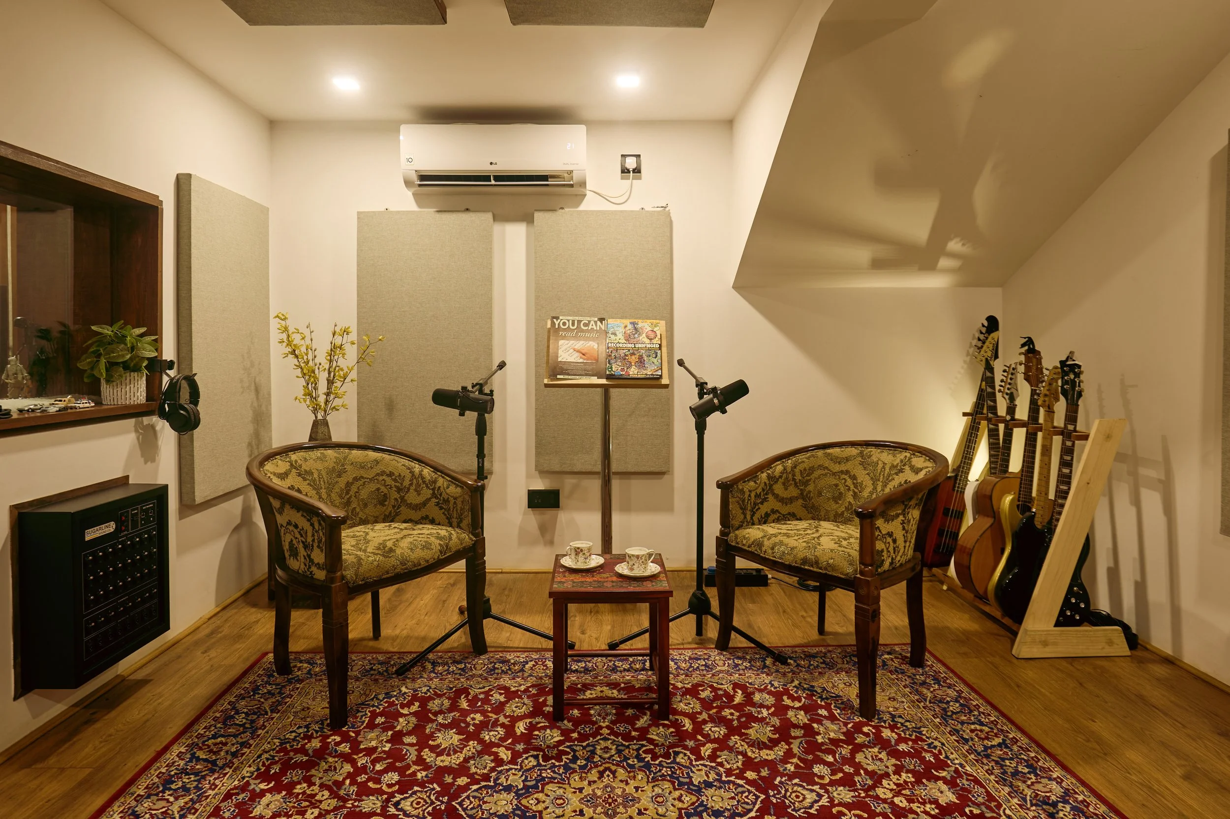Music recording studio with two vintage armchairs, microphones, a small table with cups, guitars on a stand, and soundproofing panels on the wall.