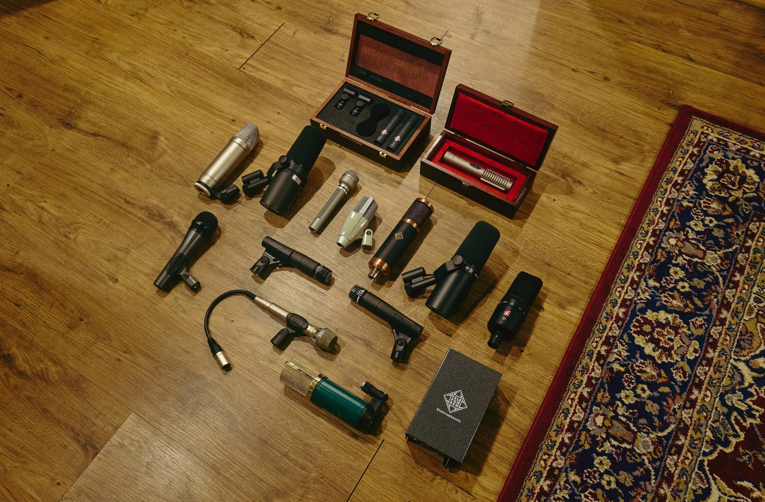 Various microphones, microphone accessories, and cases arranged on a wooden floor next to a decorative rug.