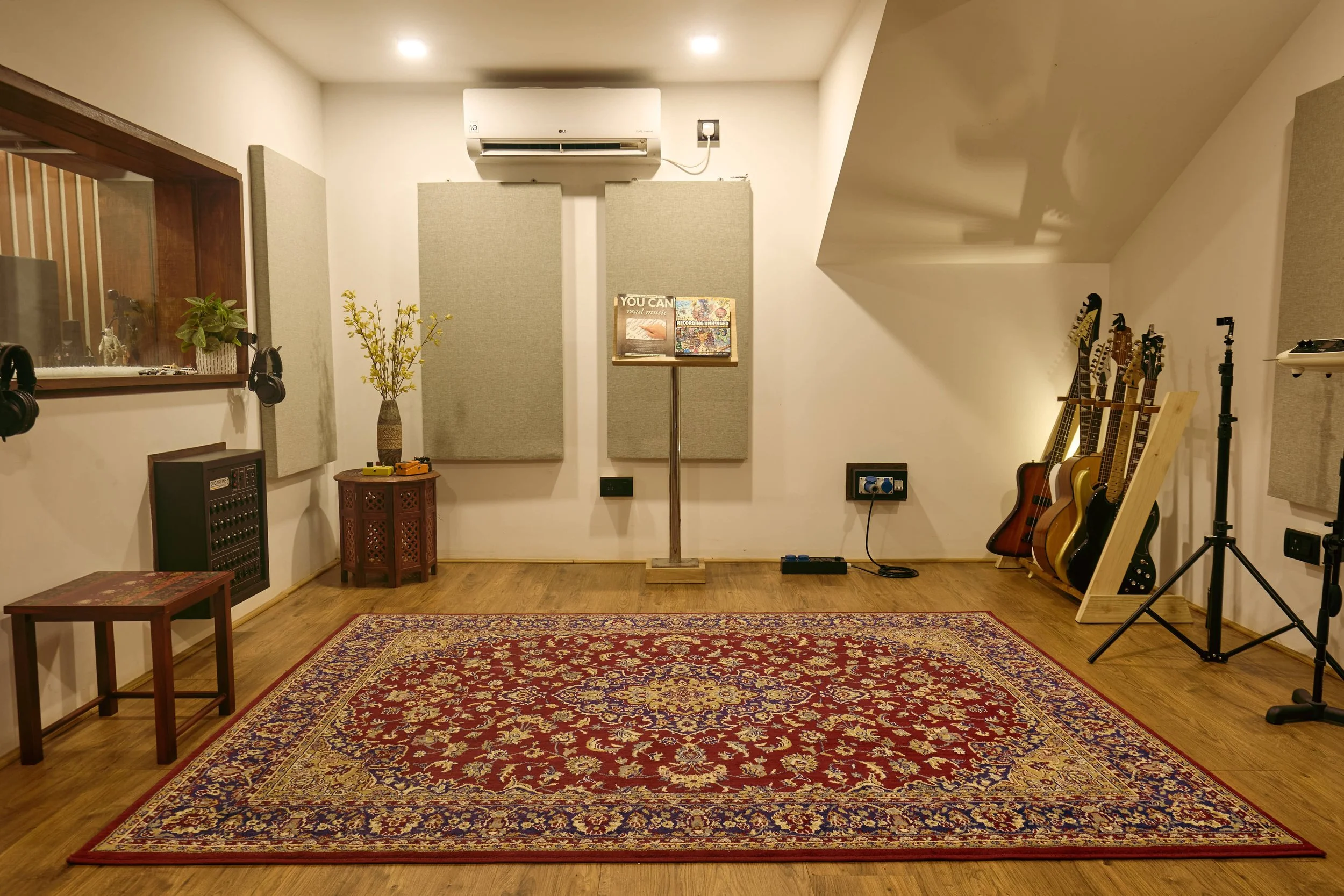 Music studio or rehearsal room with guitars on a stand, recording equipment, acoustic panels on the walls, a large red patterned rug on wooden floor, and a window looking into a recording booth.