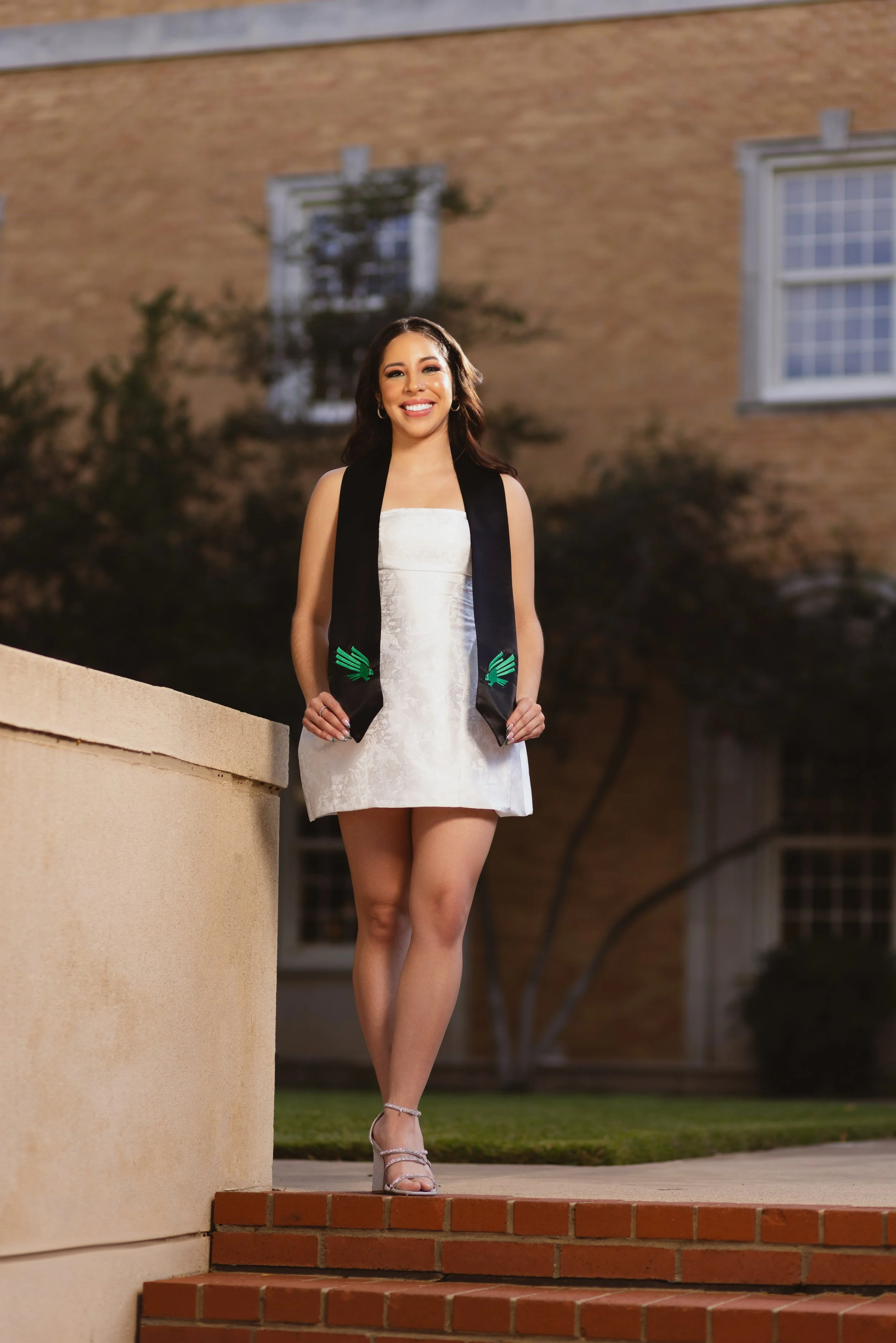 A young woman in a white dress and high heels stands on stairs outdoors at night, smiling, with a graduation stole draped around her neck.