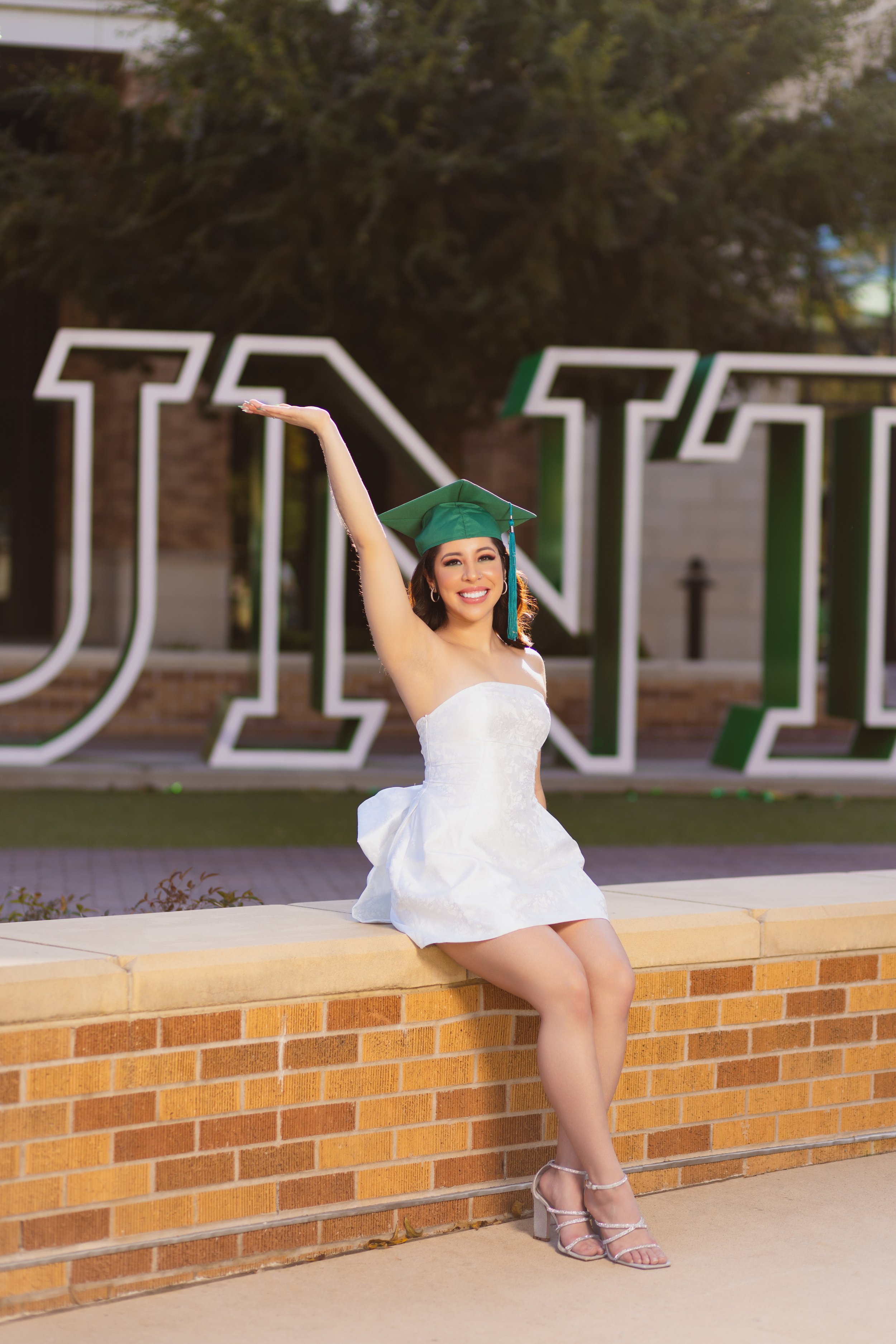 A young woman in a white dress and high heels sitting on a brick ledge, wearing a green graduation cap, smiling and posing with one arm raised outdoors, with large illuminated letters in the background.
