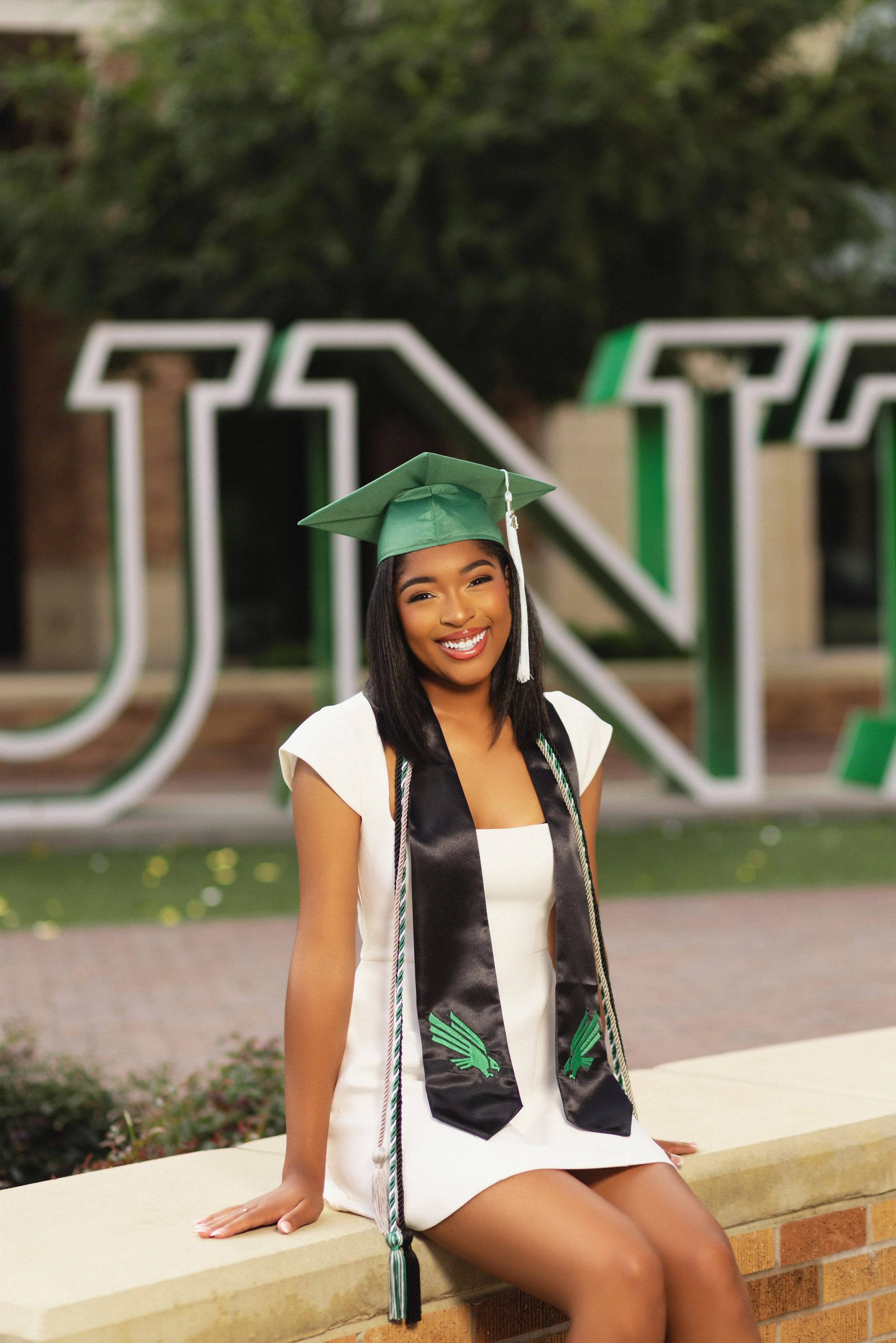 A young woman in a white dress and graduation cap, smiling and sitting on a stone wall, with a large green and white university sign in the background.