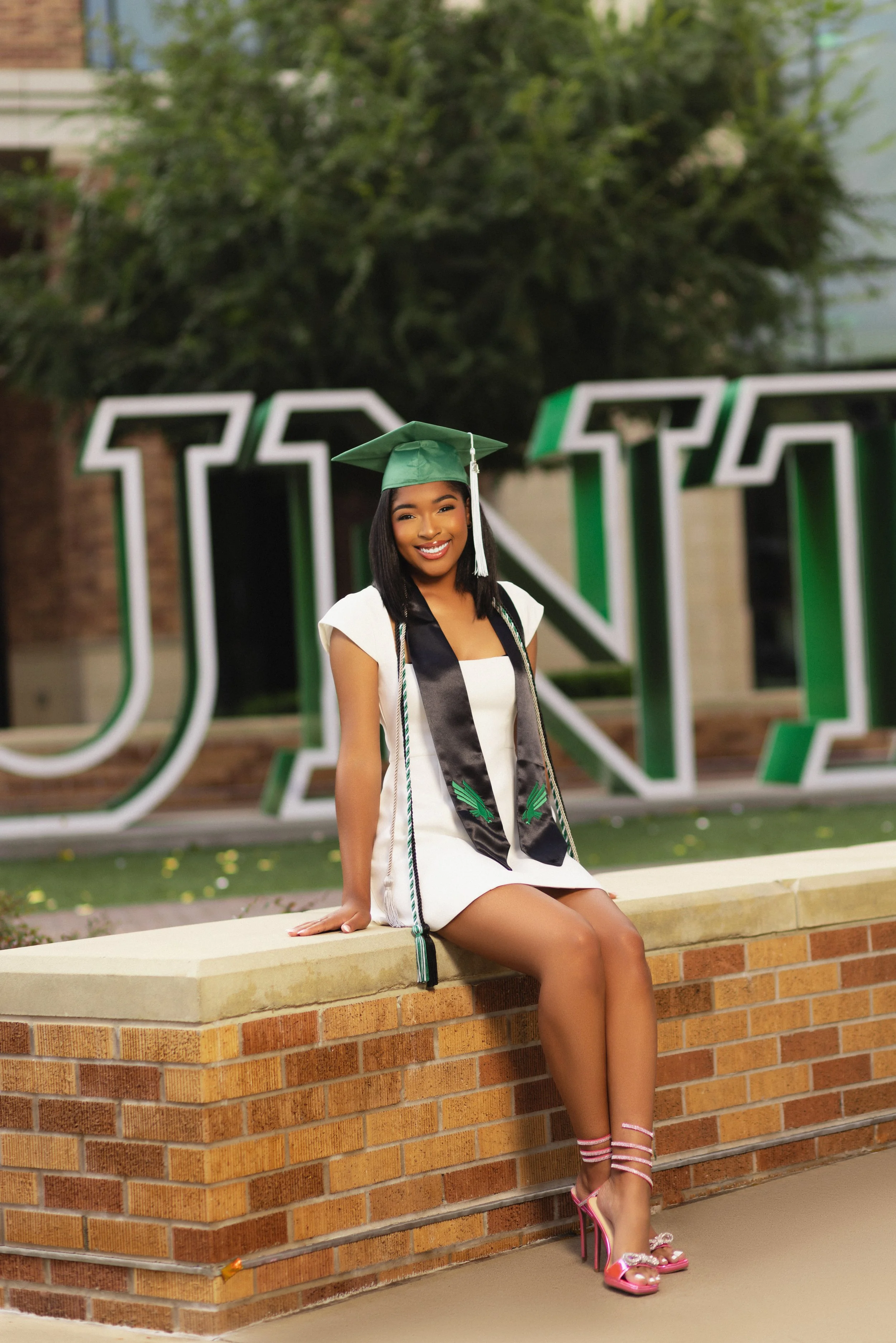 A young woman in a white dress and pink high heels sitting on a brick ledge, wearing a green graduation cap and gown sash, smiling at the camera, with large white and green letters in the background.