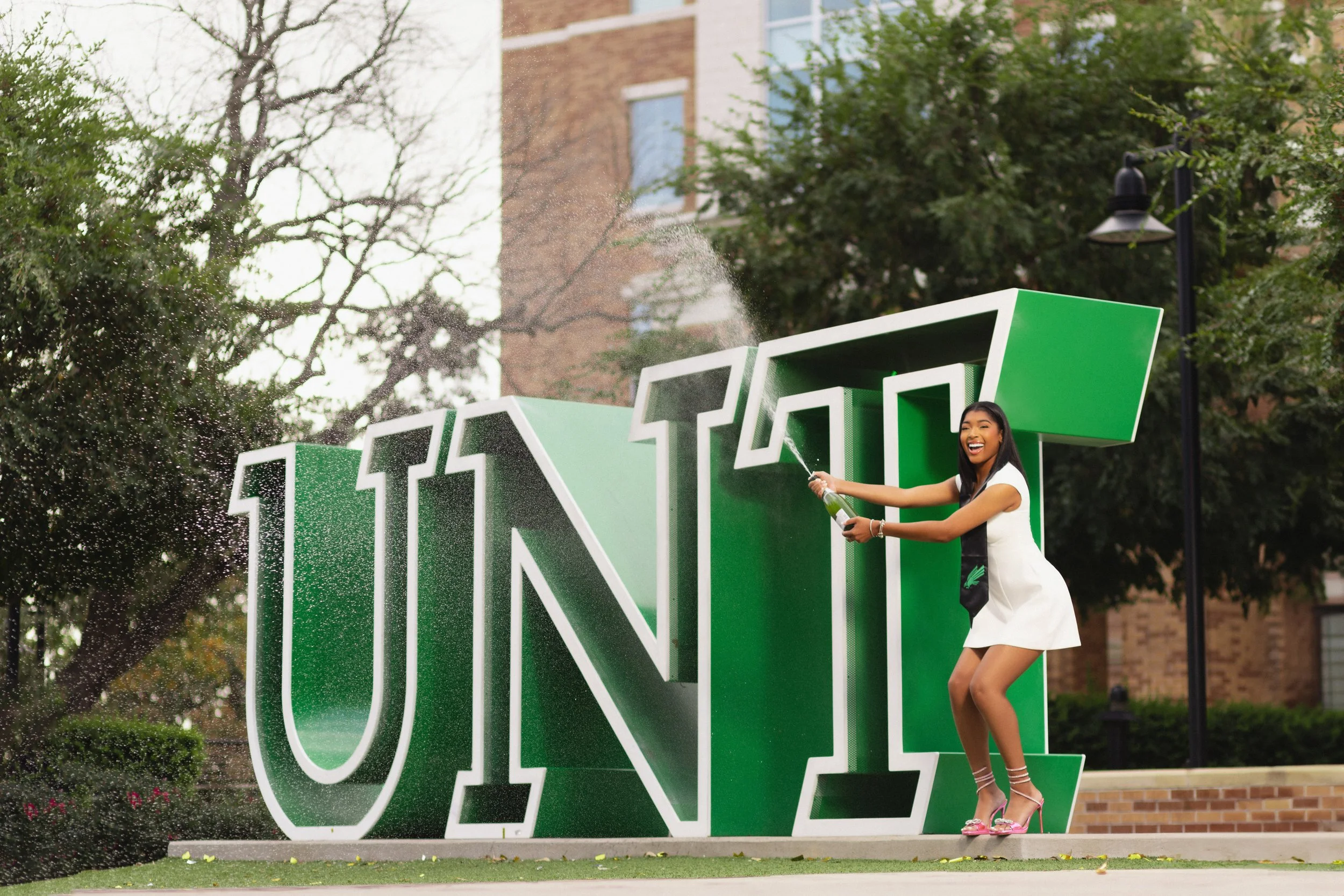 A young woman in a white dress and high heels is celebrating on a university campus, standing next to a large green and white sign that reads 'U N I T' while spraying champagne.