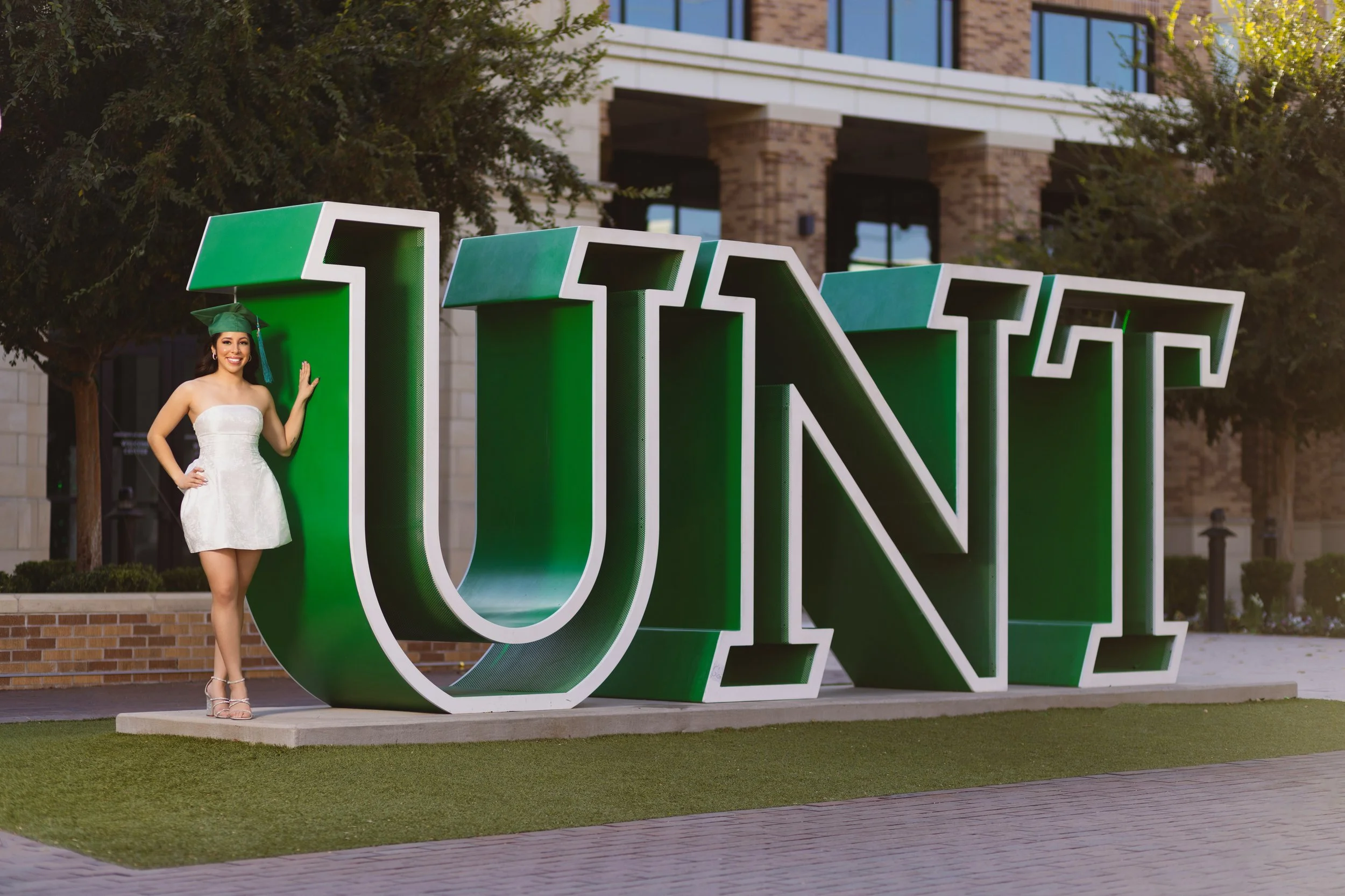 A young woman in a white dress and graduation cap standing next to a large green and white 'UNIVERSITY' sign outdoors.