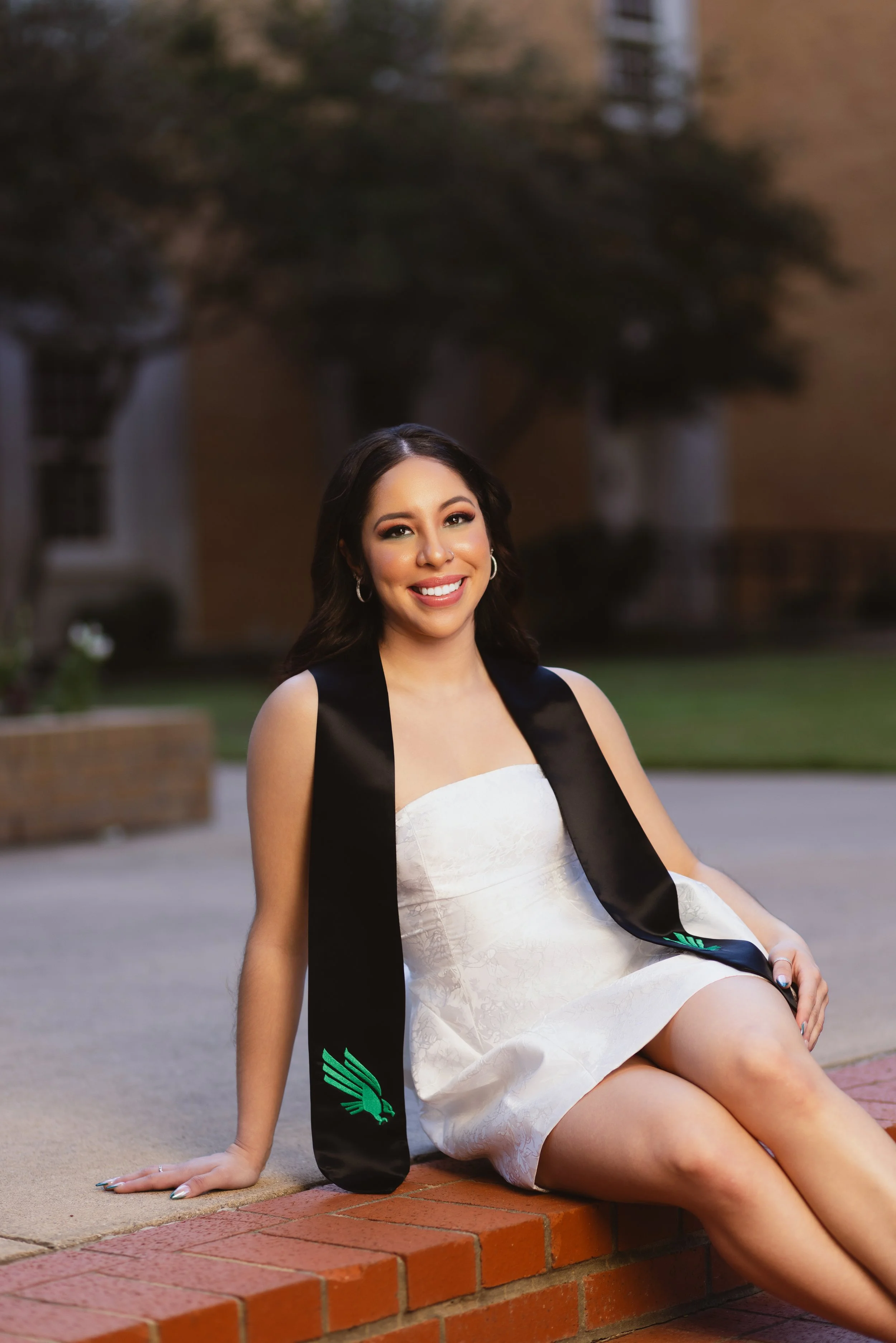 A young woman smiling and sitting on a brick ledge outdoors, wearing a white dress and a black sash, with a graduation cap in hand.