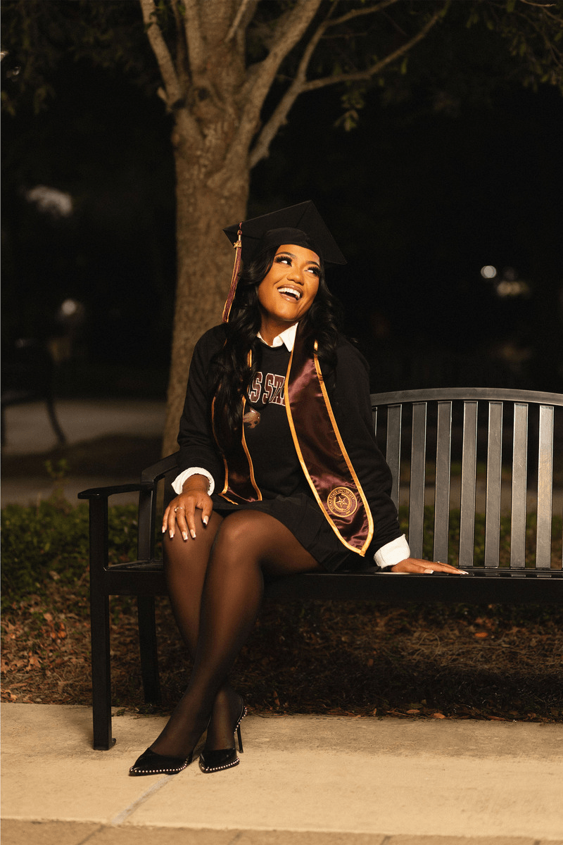 A young woman in a graduation cap and gown with a stole, sitting on a park bench at night, smiling and looking up.